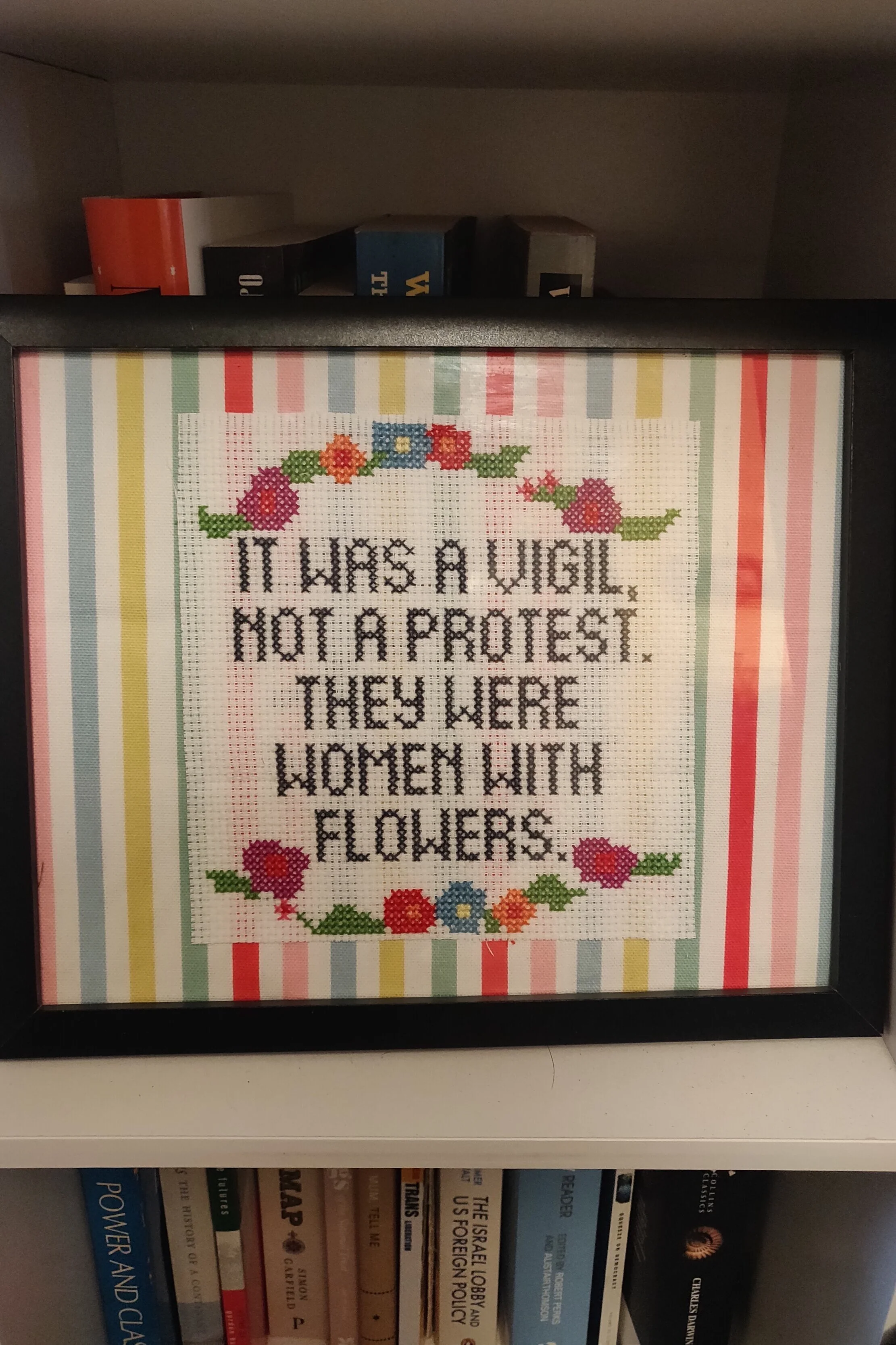 A framed cross-stitch piece leaned on a bookcase, with the phrase “It was a vigil, not a protest. They were women with flowers” surrounded by blue, pink, and orange flowers.