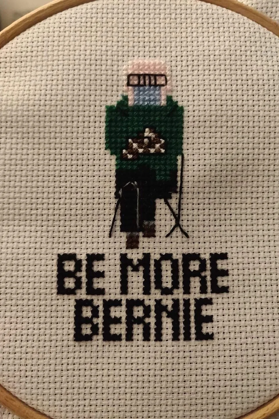 Close-up of a cross-stitched version of the viral photo of Bernie Sanders sitting in a mask and mittens at the Biden inauguration, with the phrase “Be More Bernie” below.