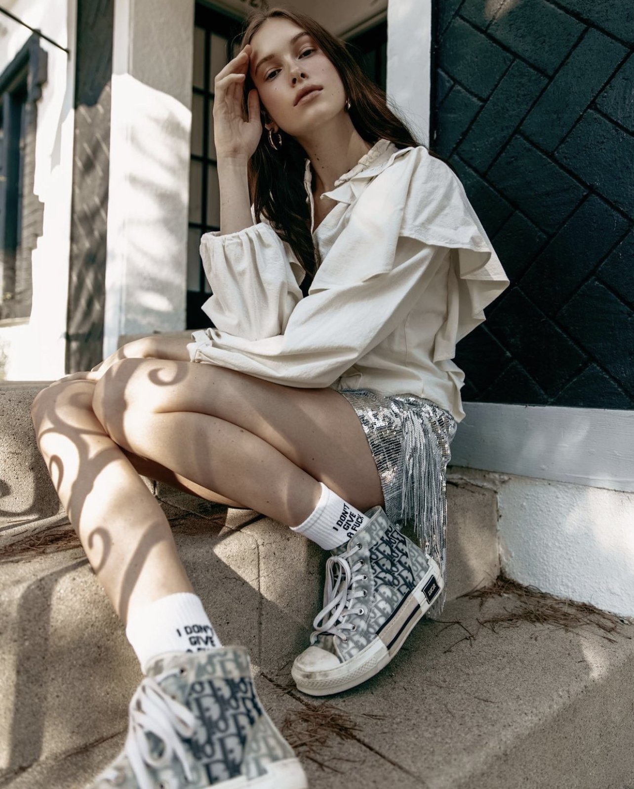 Young woman sitting on steps outside, wearing a white blouse, shiny silver shorts, high-top sneakers, and white socks with black text, with shadows from a decorative fence cast on her legs.