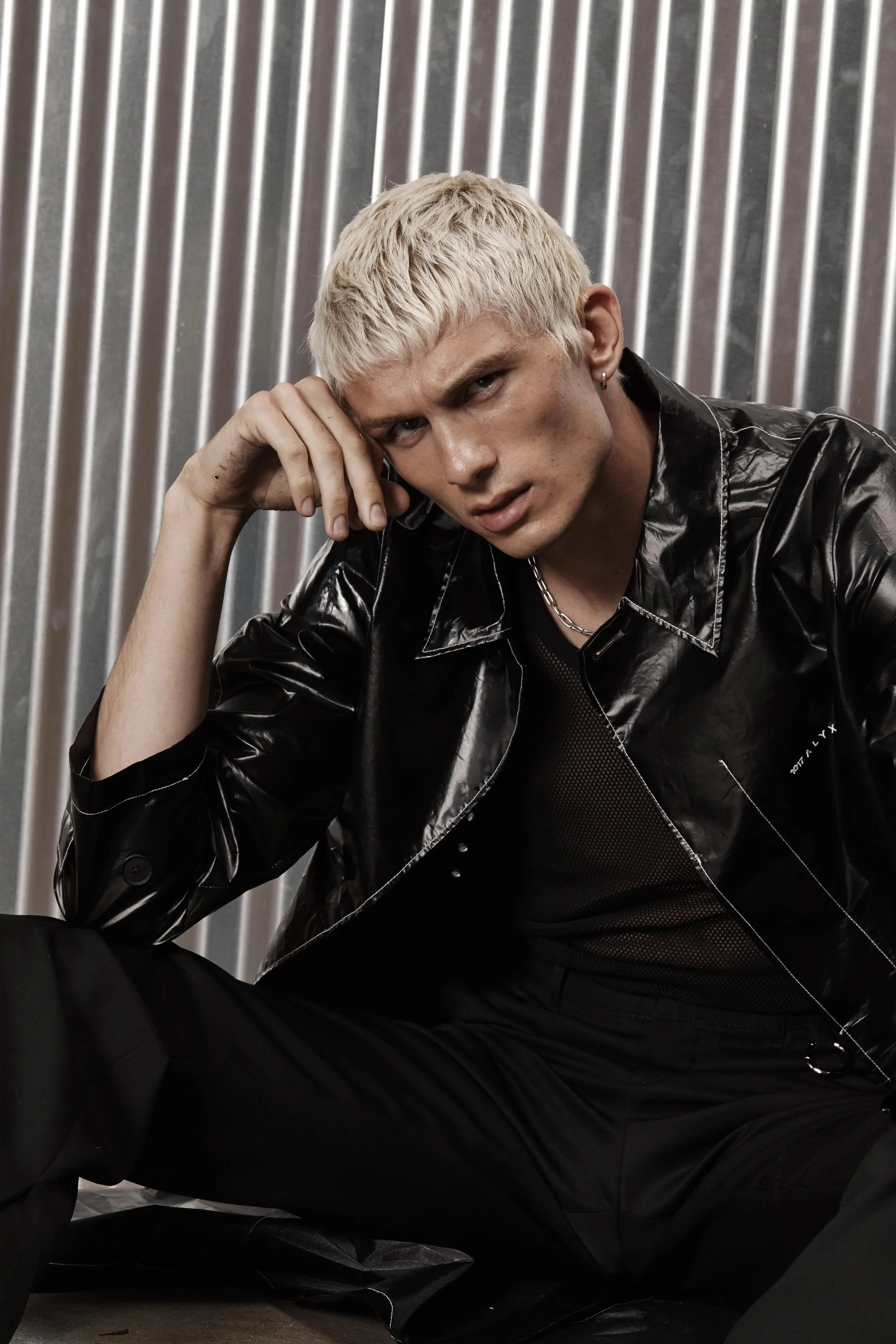 A black and white photo of a young man with platinum blonde hair sitting against a metallic striped background. He is wearing a shiny black leather jacket, black shirt, and a chain necklace, with a serious expression and resting his head on his hand.