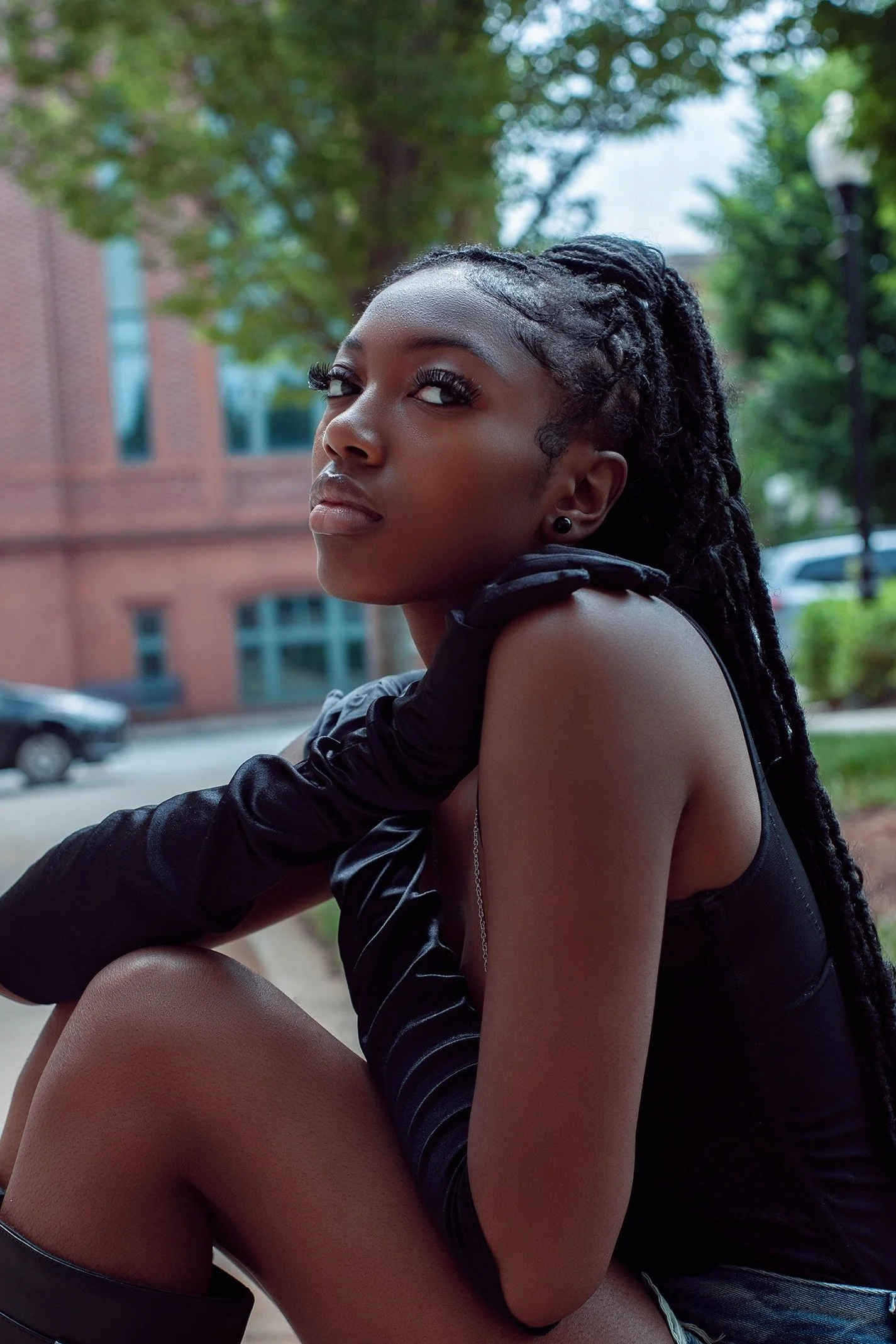 A young Black woman with dreadlocks sitting outdoors on a city street, wearing a black sleeveless top and long black glove on her left arm, looking thoughtfully at the camera with trees and a building in the background.