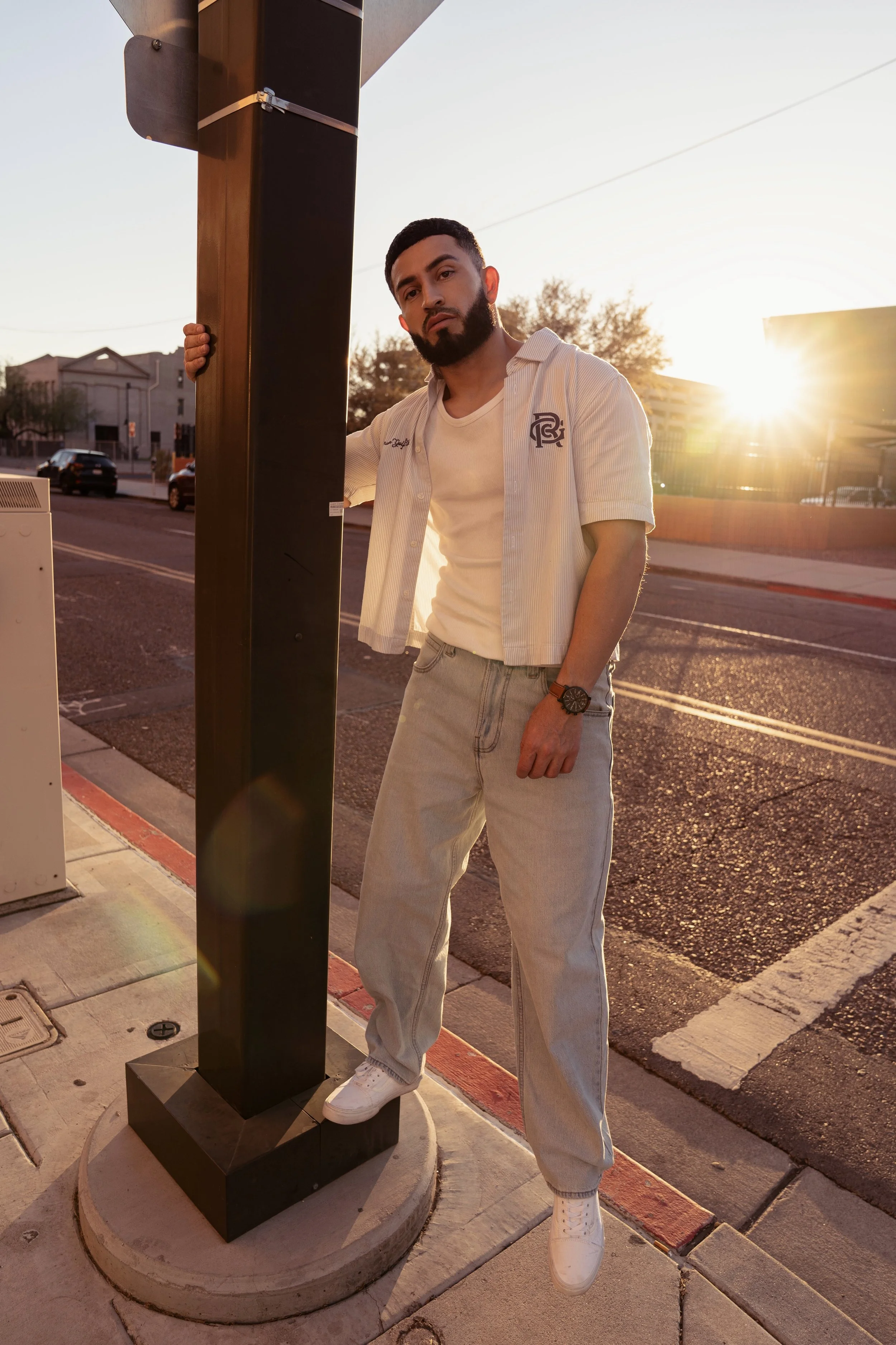A young man with a beard stands on a city sidewalk at sunset, wearing a beige t-shirt, an open button-up shirt, light jeans, and white sneakers. He leans on a black pole, gazing at the camera with a serious expression, with the sun setting behind him