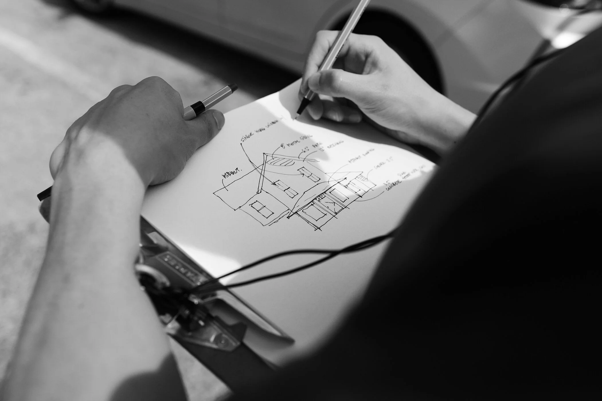 Close-up of an architect's hands sketching a house design on a clipboard outdoors. The drawing features detailed annotations and a car is visible in the background
