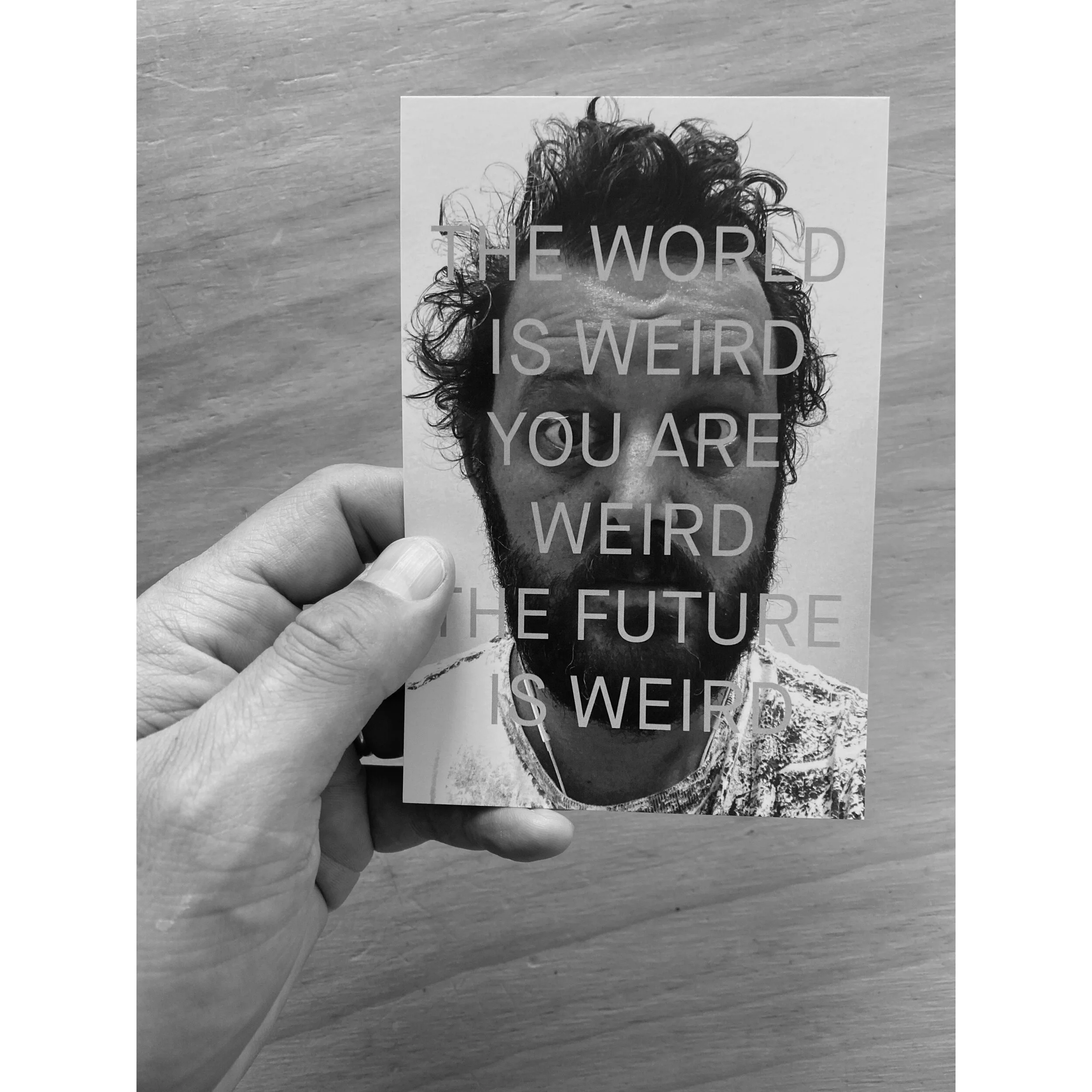 Brion Nuda Rosch holds a postcard with the text THE WORLD IS WEIRD YOU ARE THE WEIRD THE FUTURE IS WEIRD over a picture of the artist
