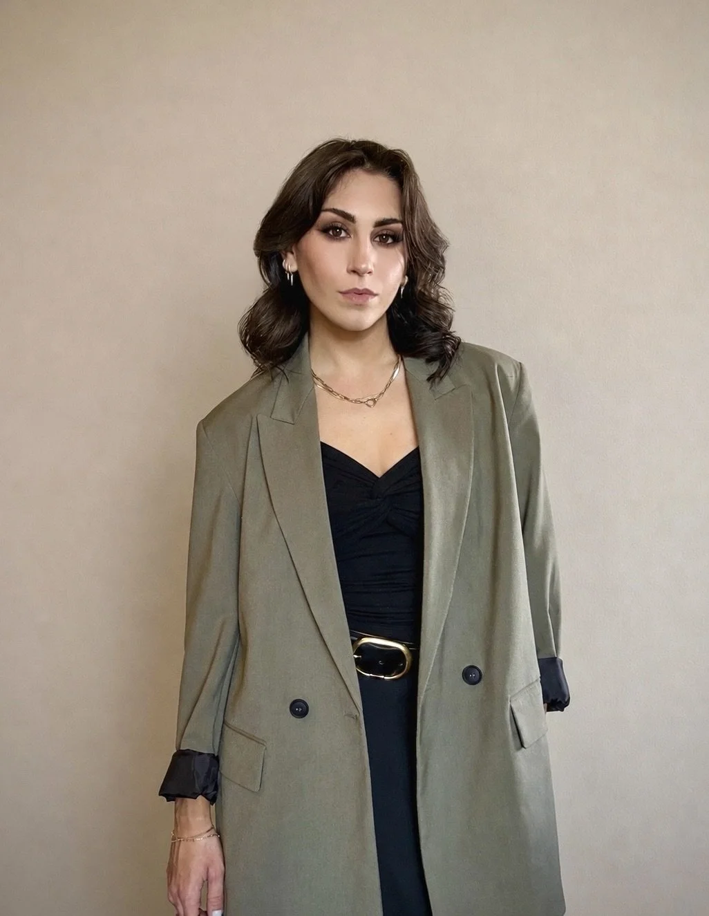 dark brunette woman with shoulder length hair and dark brown eyes in makeup with gold jewelry, a black top, black skirt, gold and black belt, and an olive double breasted blazer