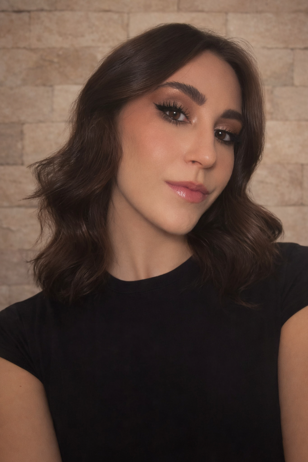 brunette women with warm smokey eye and dusty rose lips in black tshirt against warm stone brick wall