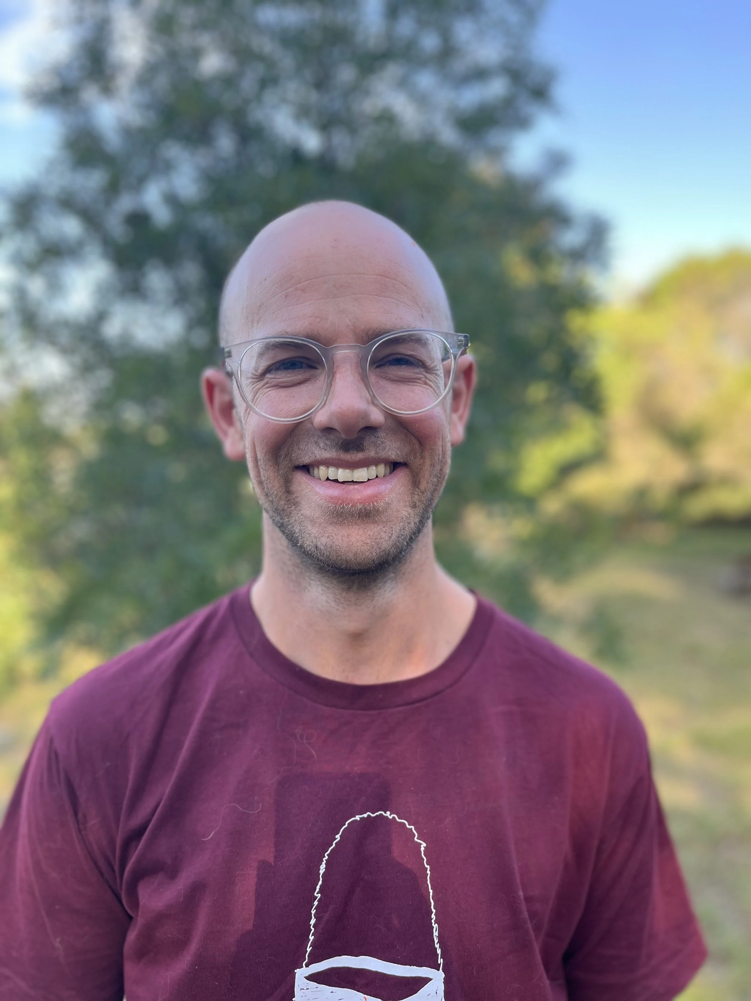 Jem – A smiling male presenting non-binary person with glasses standing outdoors with trees and blue sky in the background.