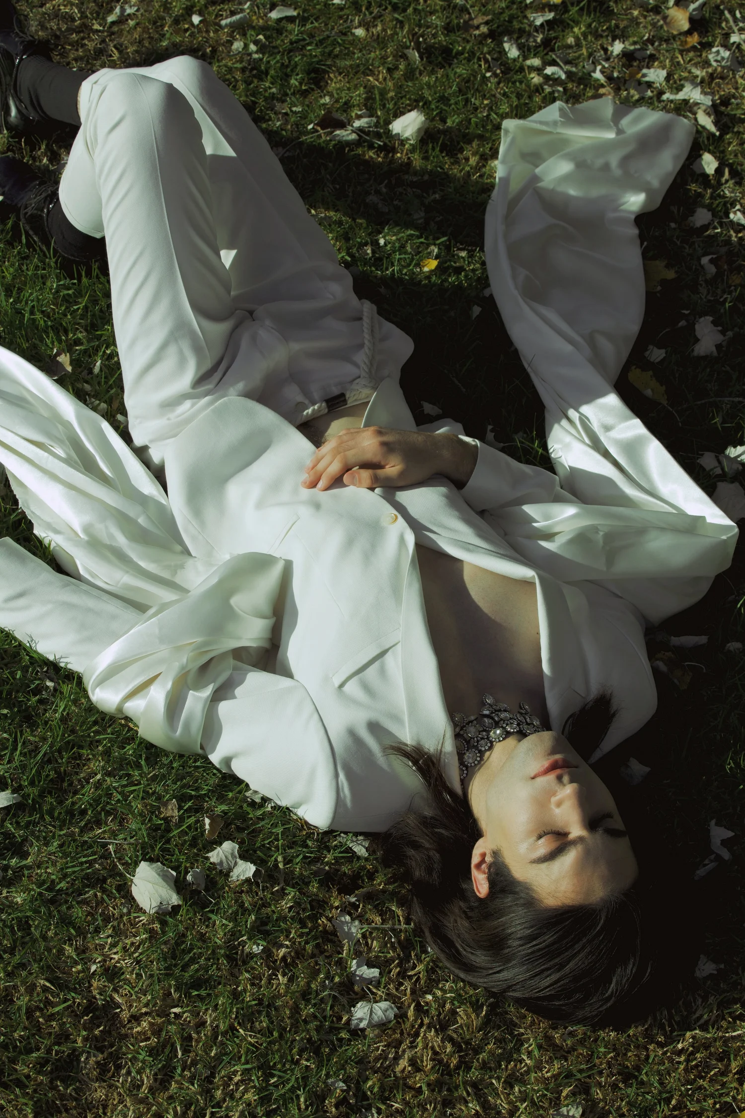 Person in a flowing white outfit lying on grass with eyes closed and jeweled face adornment