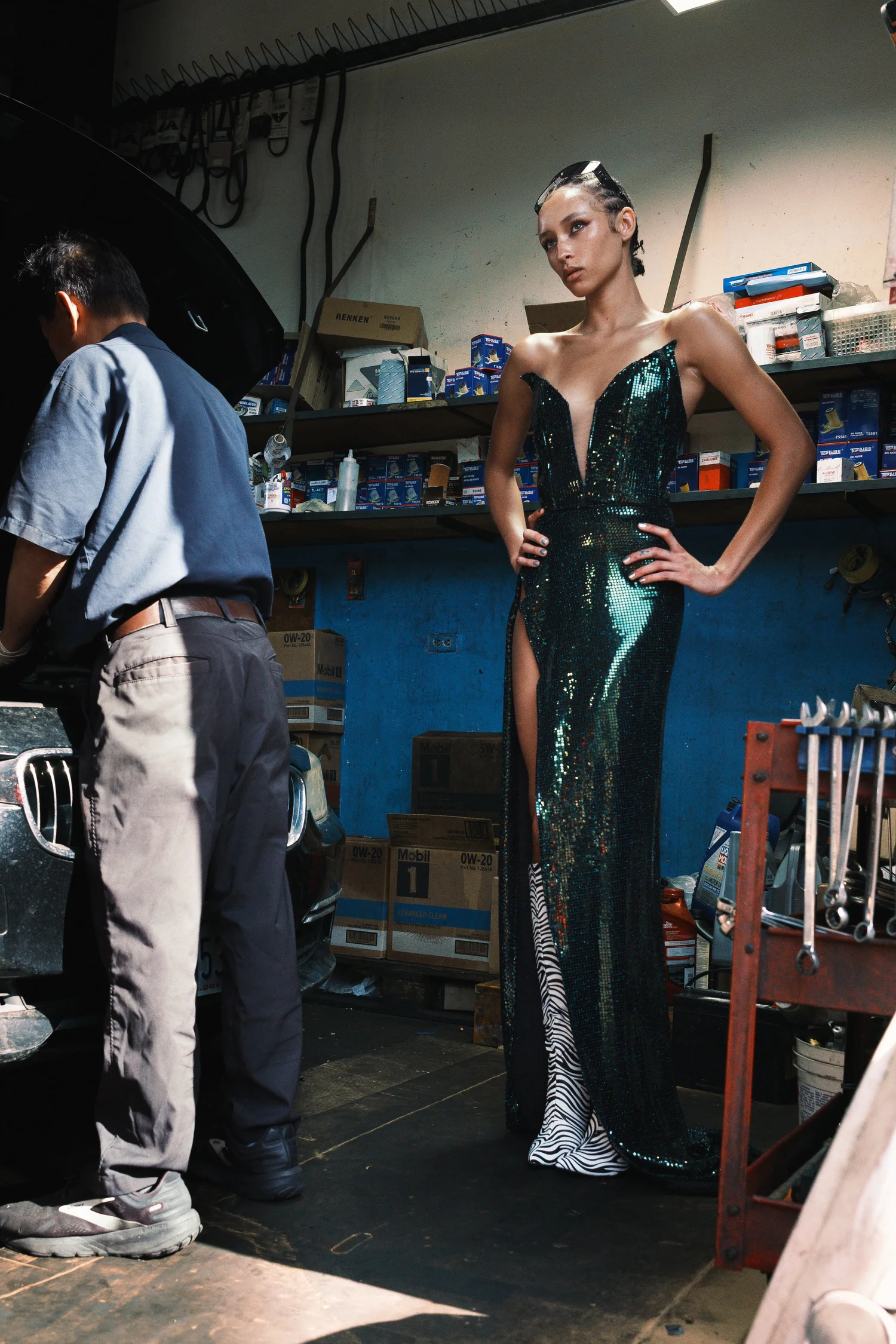 Model in a black sequined dress and sunglasses poses beside a mechanic in an auto repair shop stocked with oil filters and supplies.