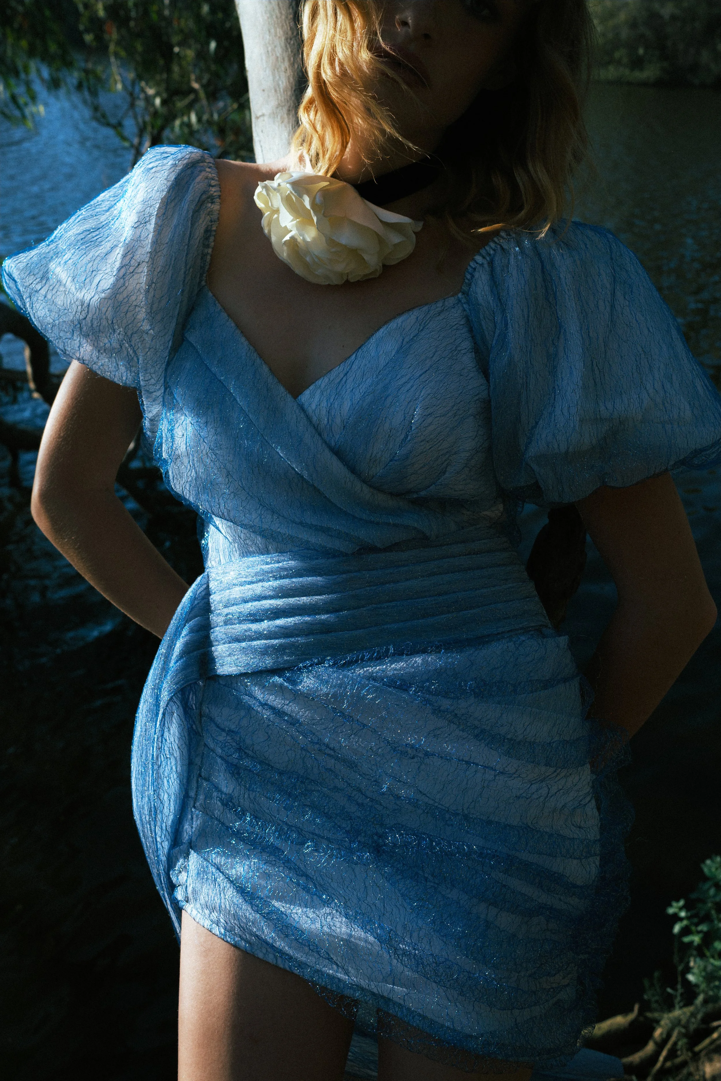 Close-up of a woman in a sheer blue textured dress with a large white flower at the neckline, posing outdoors near water.