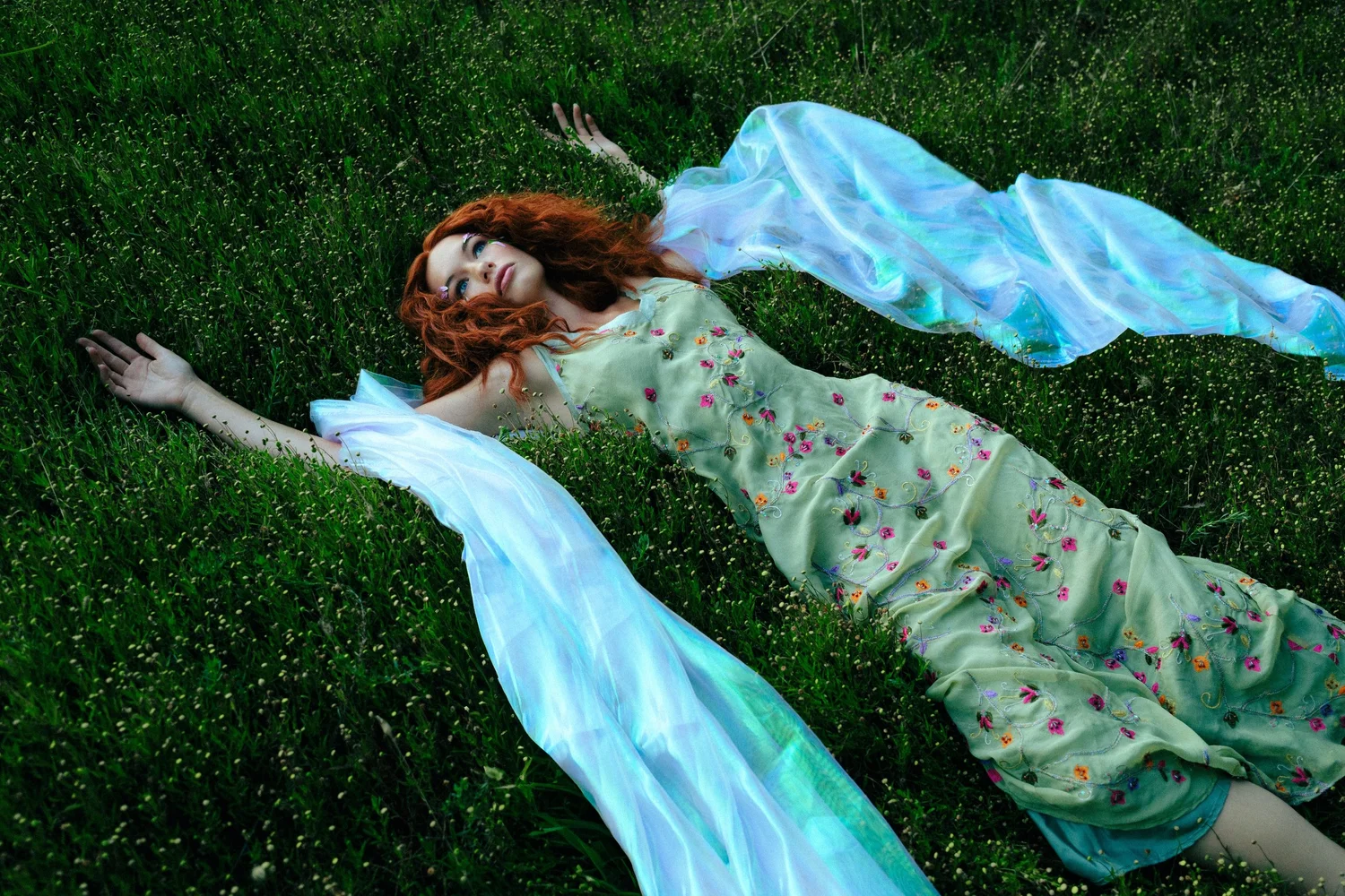 Red-haired woman lying in grass wearing a pale green floral dress with flowing translucent blue sleeves
