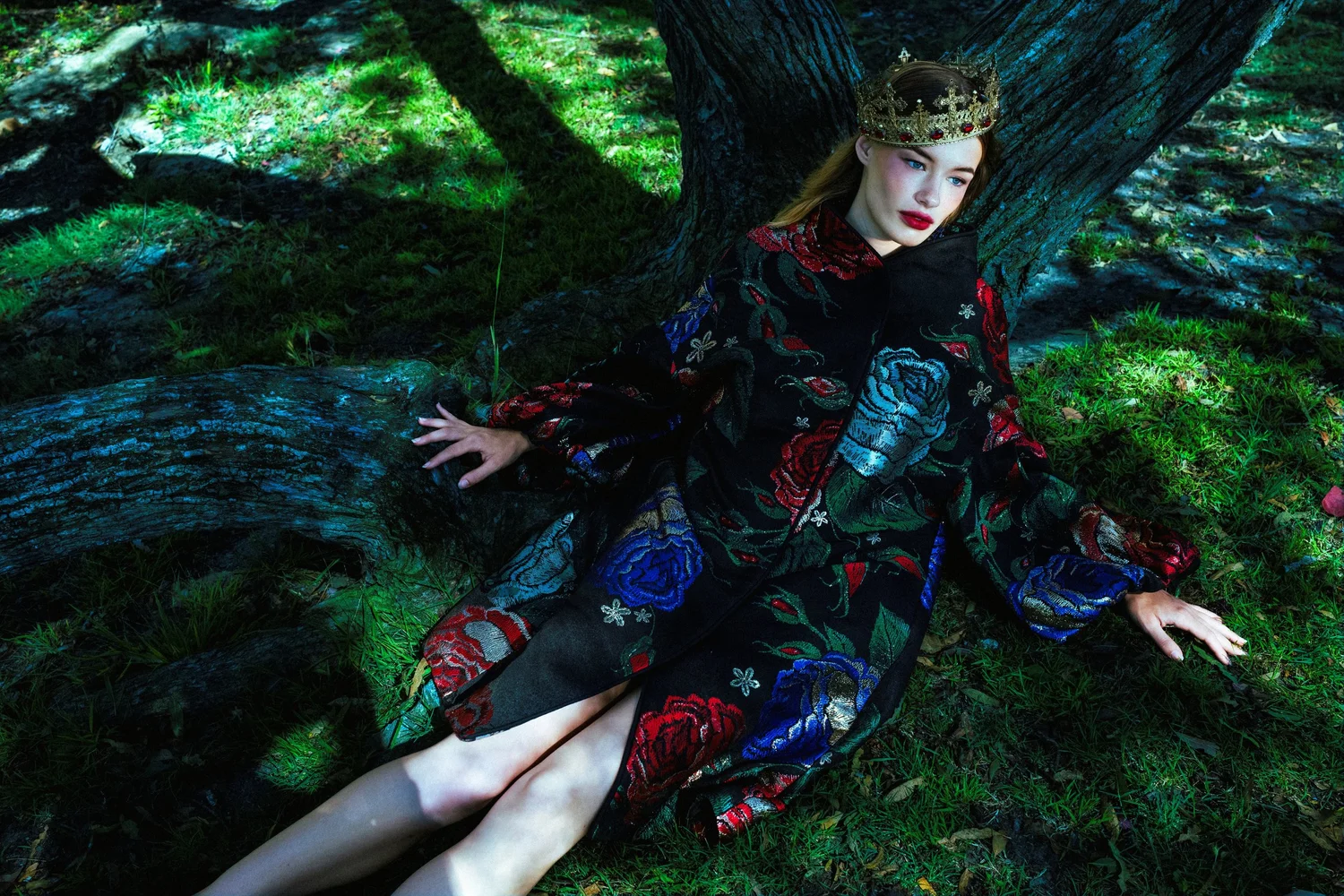 Fashion portrait of a woman wearing a jeweled crown and embroidered black floral dress, seated beneath a tree in a forest.