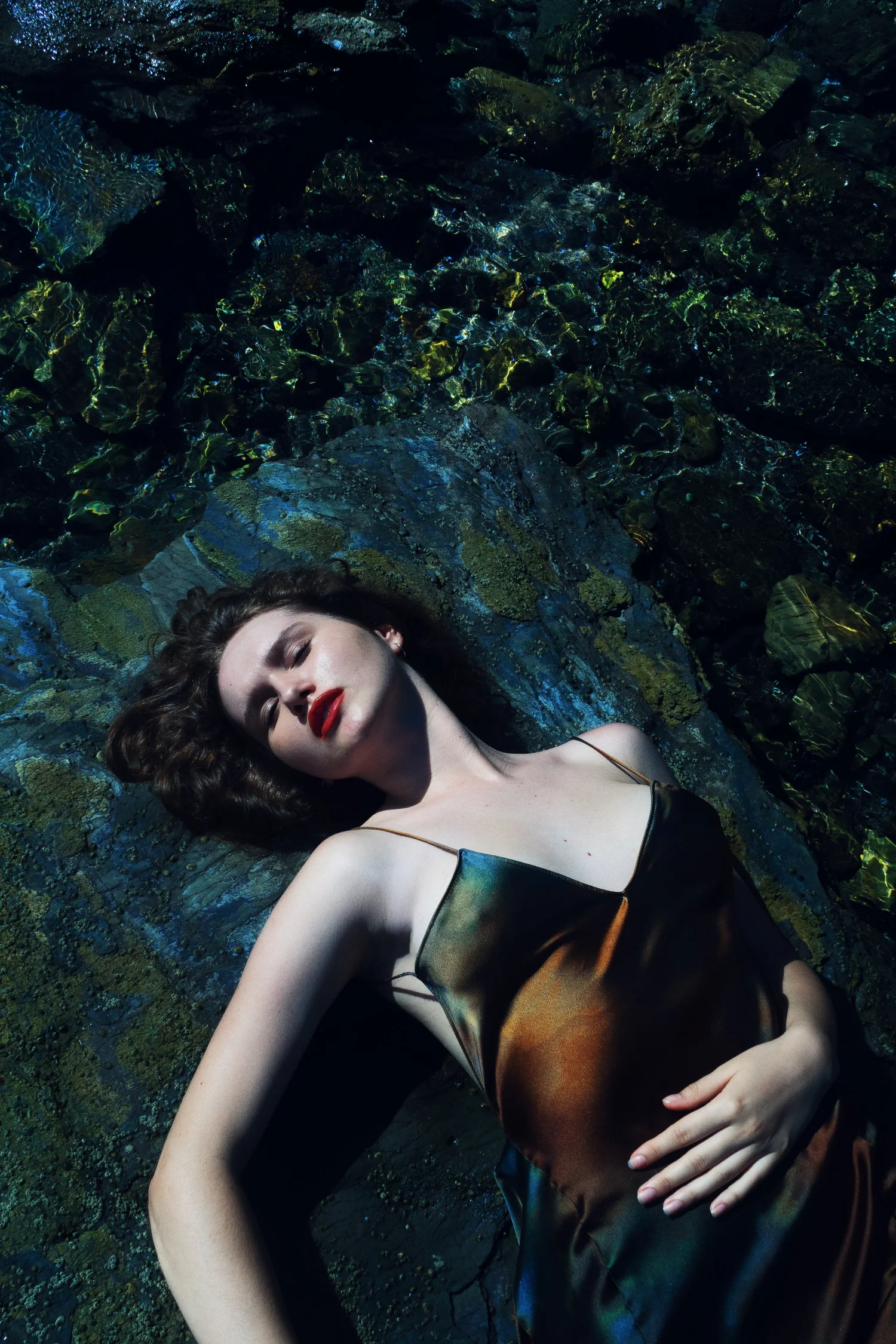 Woman with red lipstick lying on a textured, dark blue-green surface wearing a black satin dress
