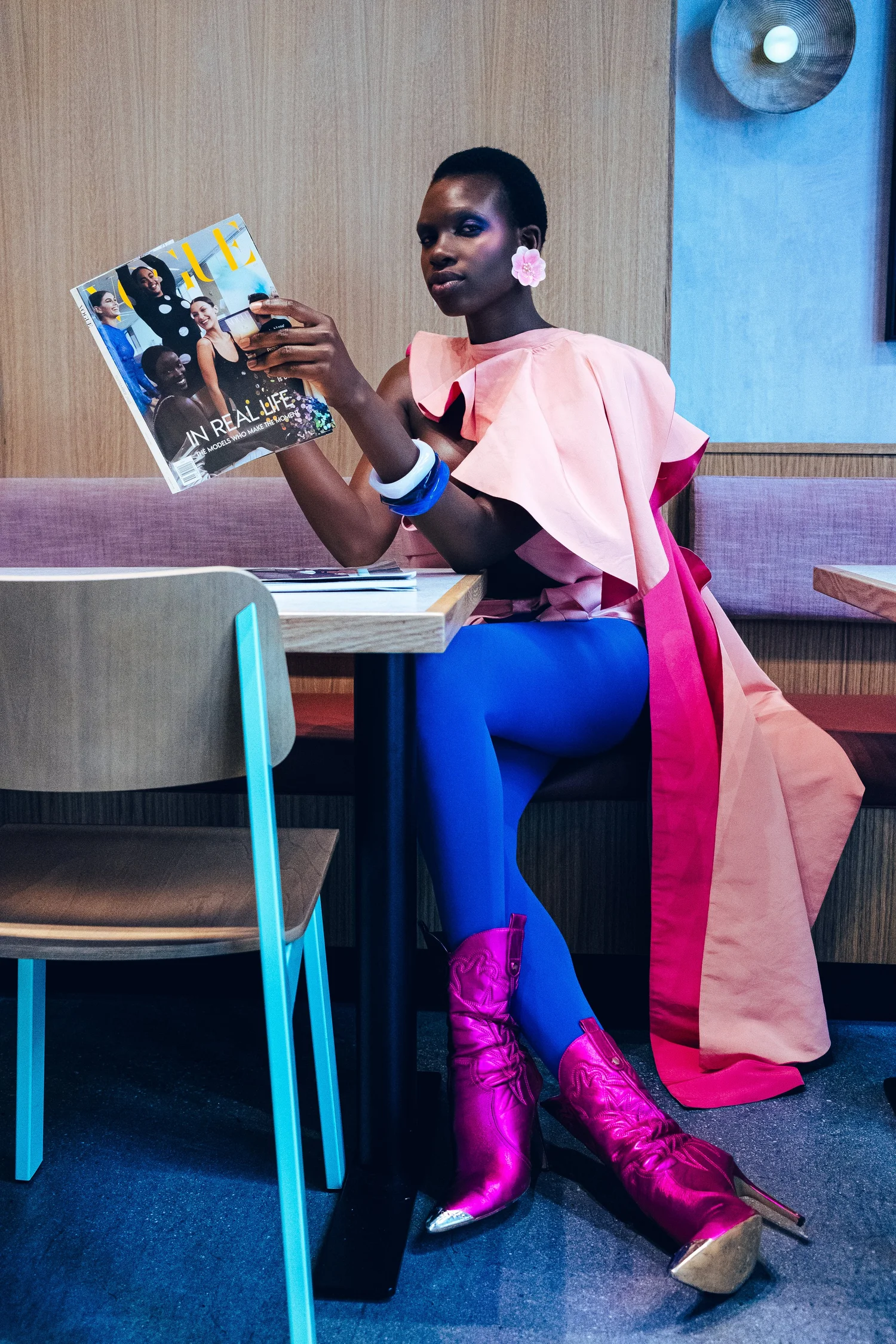A fashionably dressed Black woman in a pink ruffled top, blue pants, and metallic pink boots sits on a couch reading a fashion magazine.