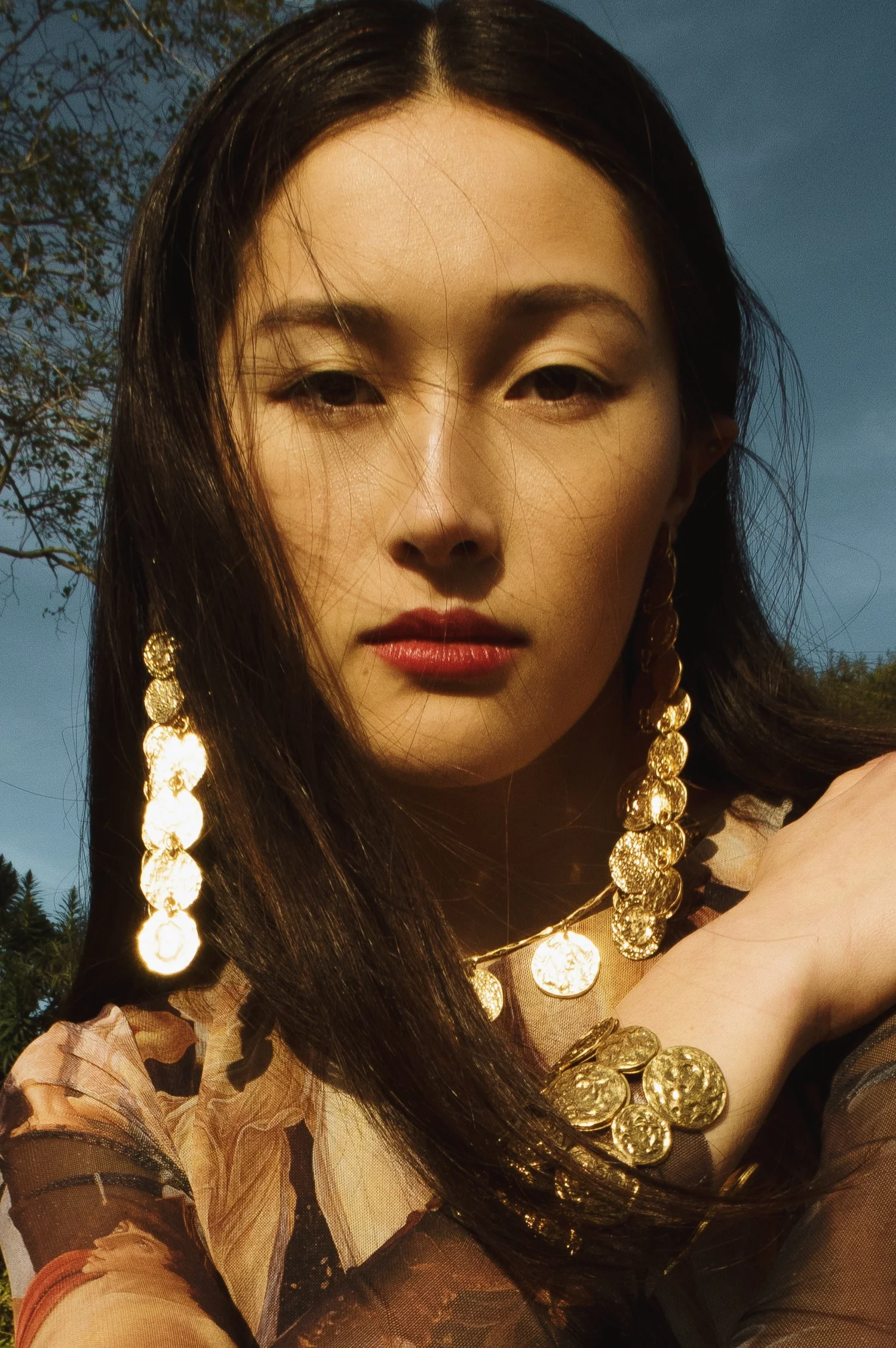 Close-up portrait of a woman with long dark hair wearing ornate gold earrings and jewelry outdoors