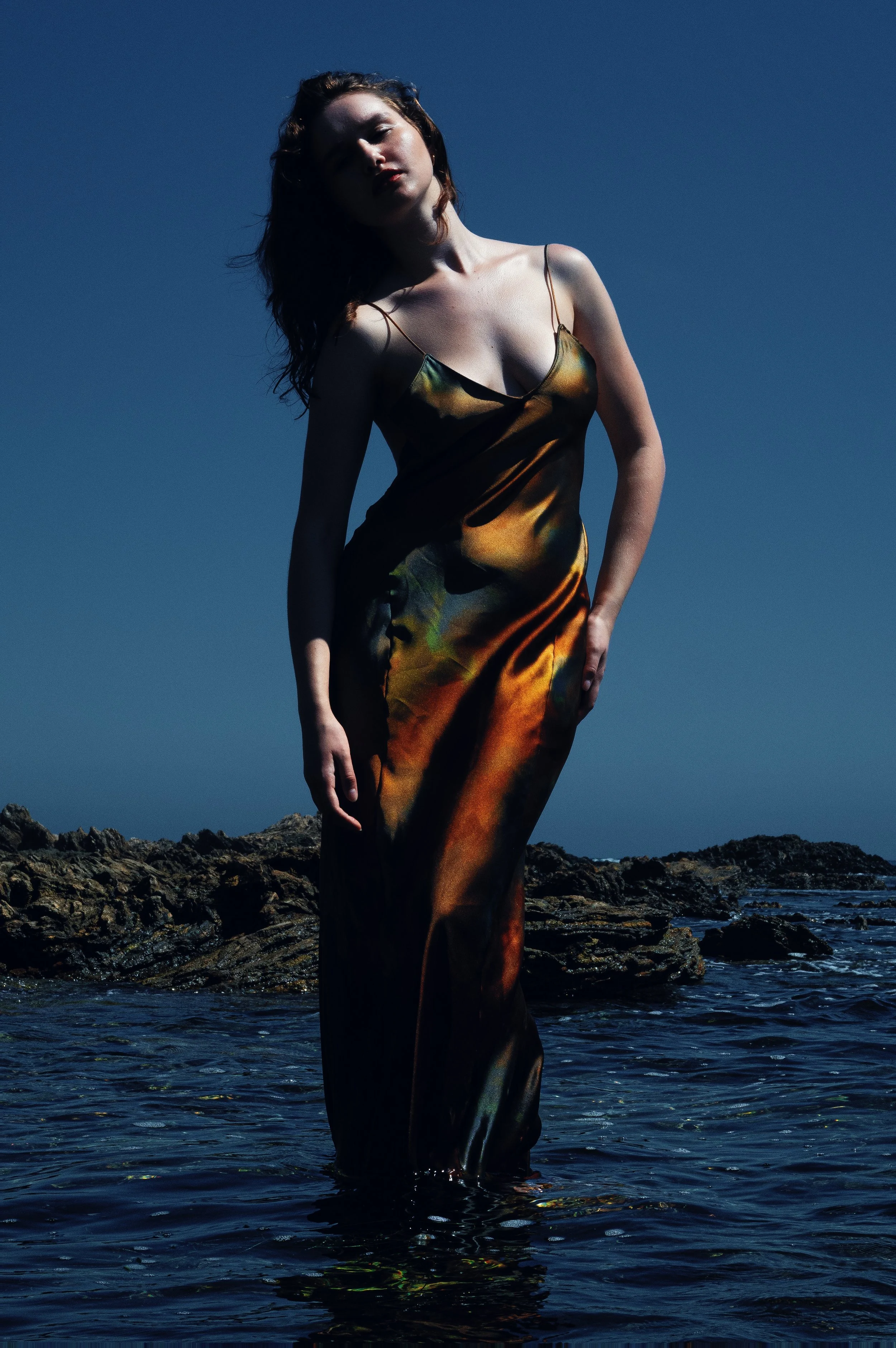 Woman in a satin slip dress standing in shallow ocean water beside a rocky shore