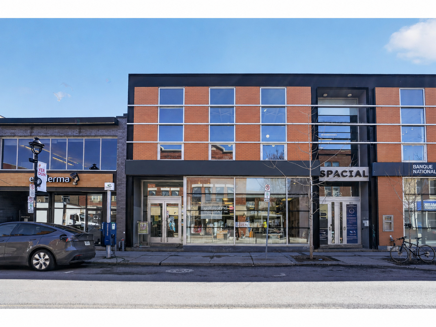 A street view of a commercial building with large glass windows. The ground floor has signs that read "Yellow" and "SPACIAL." There is a parked car and a bicycle in front of the building. The sky is blue with some clouds.