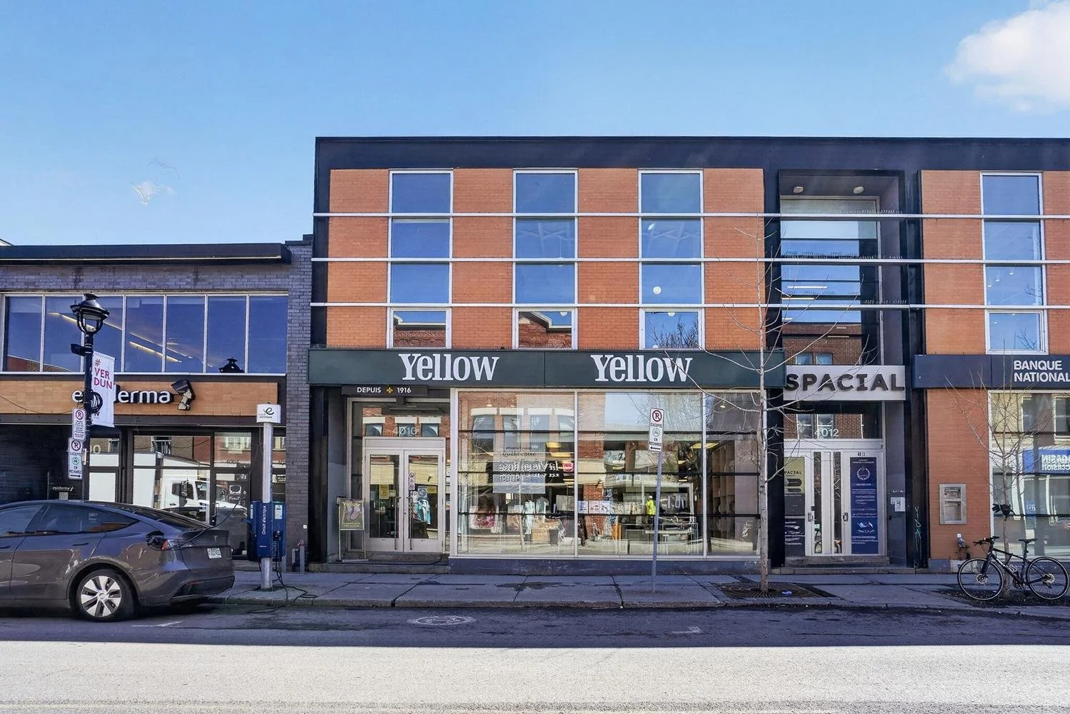 A street view of a commercial building with large glass windows. The ground floor has signs that read "Yellow" and "SPACIAL." There is a parked car and a bicycle in front of the building. The sky is blue with some clouds.