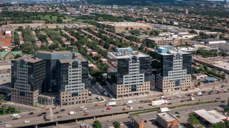 Aerial view of a city with three modern high-rise office buildings along a busy highway, with residential neighborhoods and many trees in the background.