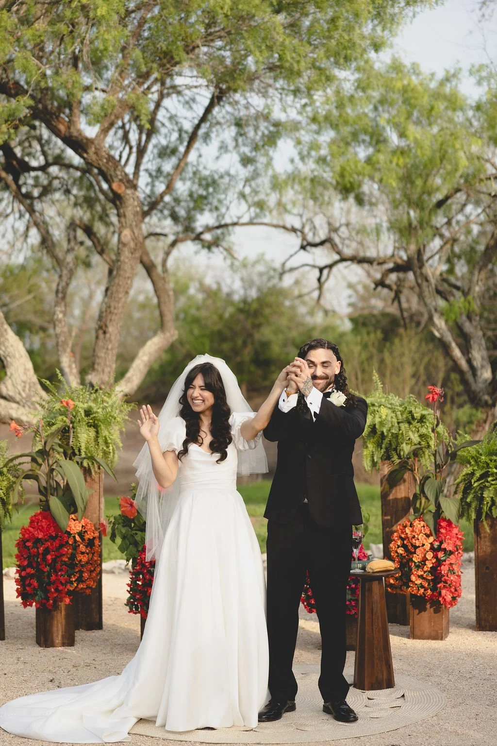 Newlywed couple celebrating at an outdoor ceremony at an Austin wedding venue, surrounded by trees, lush greenery, ferns, and vibrant red florals.