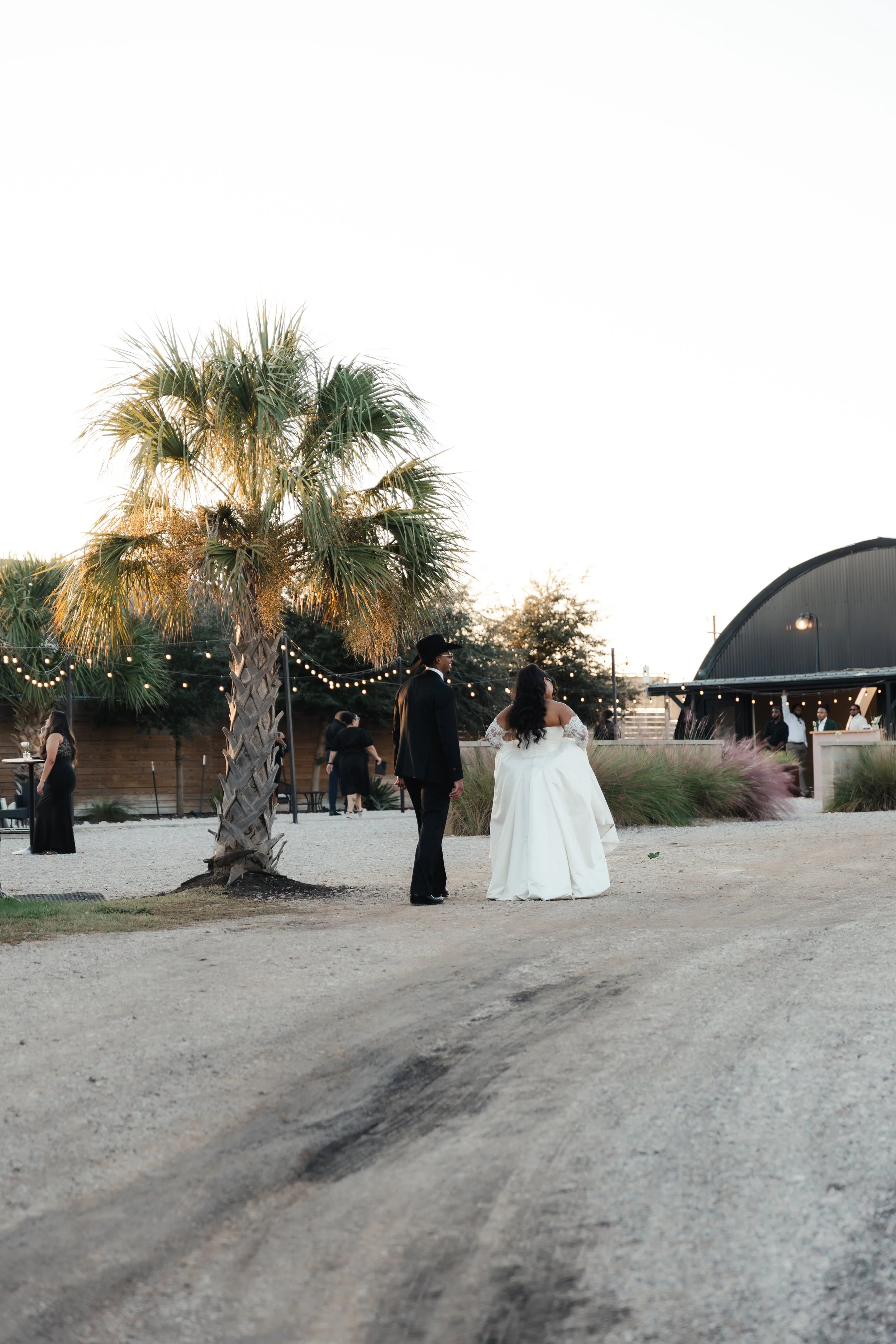 Newlywed couple walking through the courtyard at an Austin wedding venue, surrounded by palm trees, string lights, desert landscaping, and the Quonset Hut.