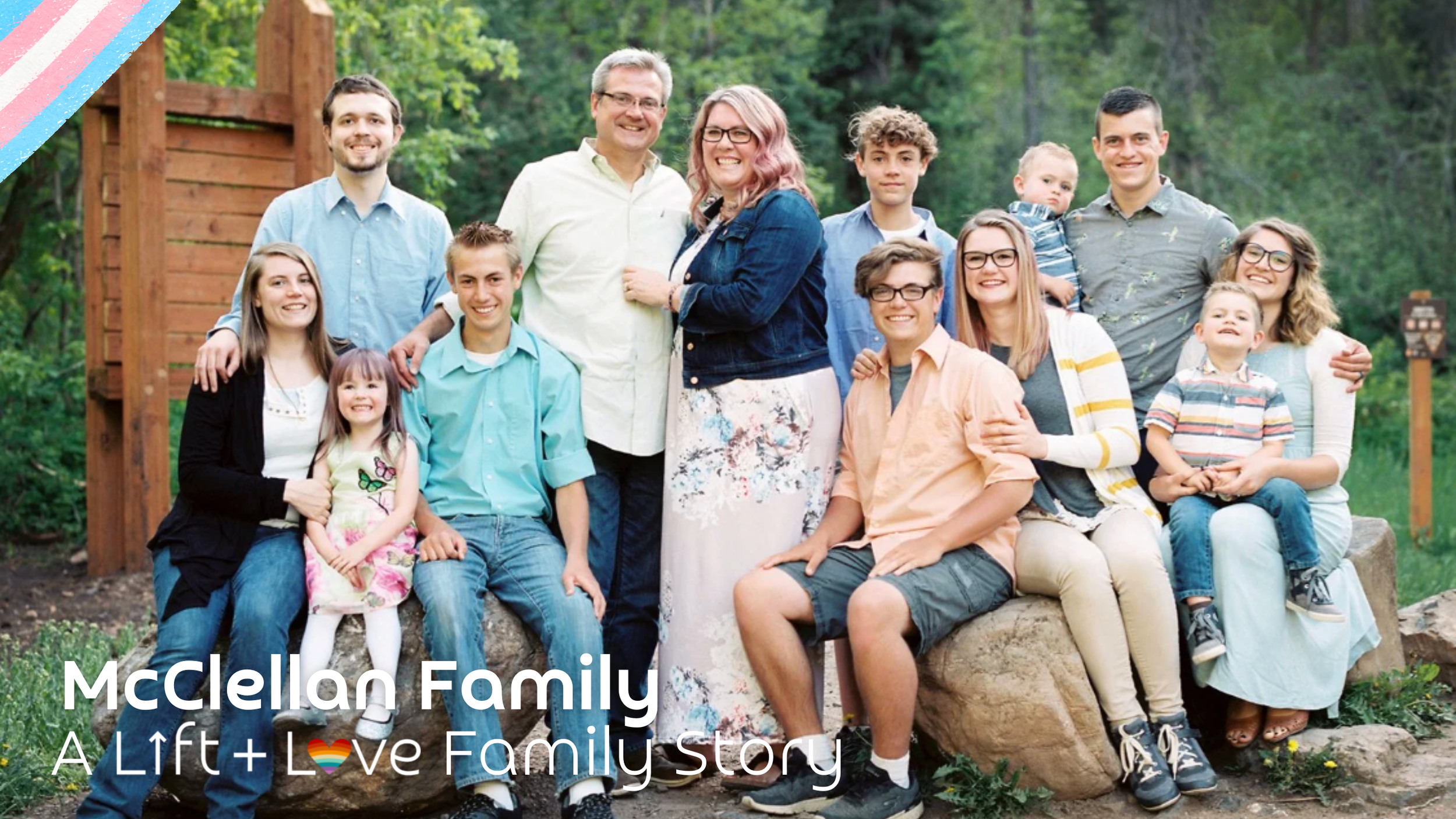A large family group of 13 people, including children and adults, posing outdoors in a green, wooded area with a wooden structure in the background, smiling for the camera.