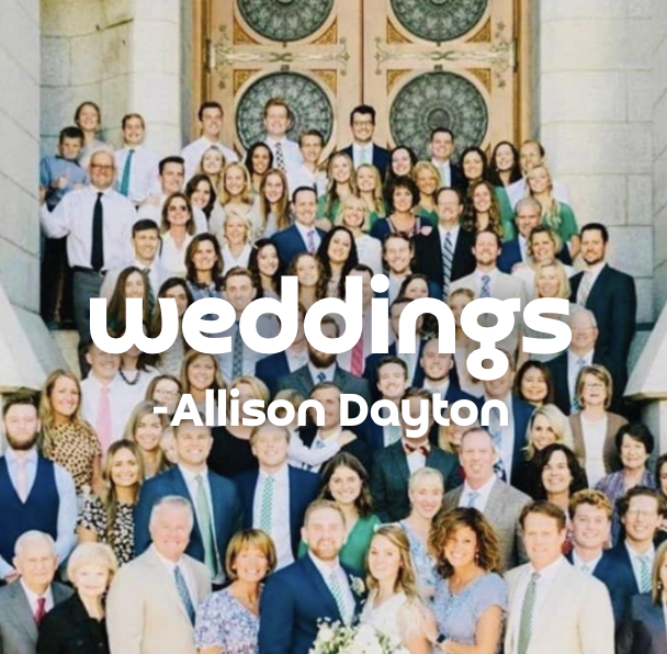 A large group of wedding guests gathered on the steps of a grand building with large decorative clocks in the background, celebrating a wedding.