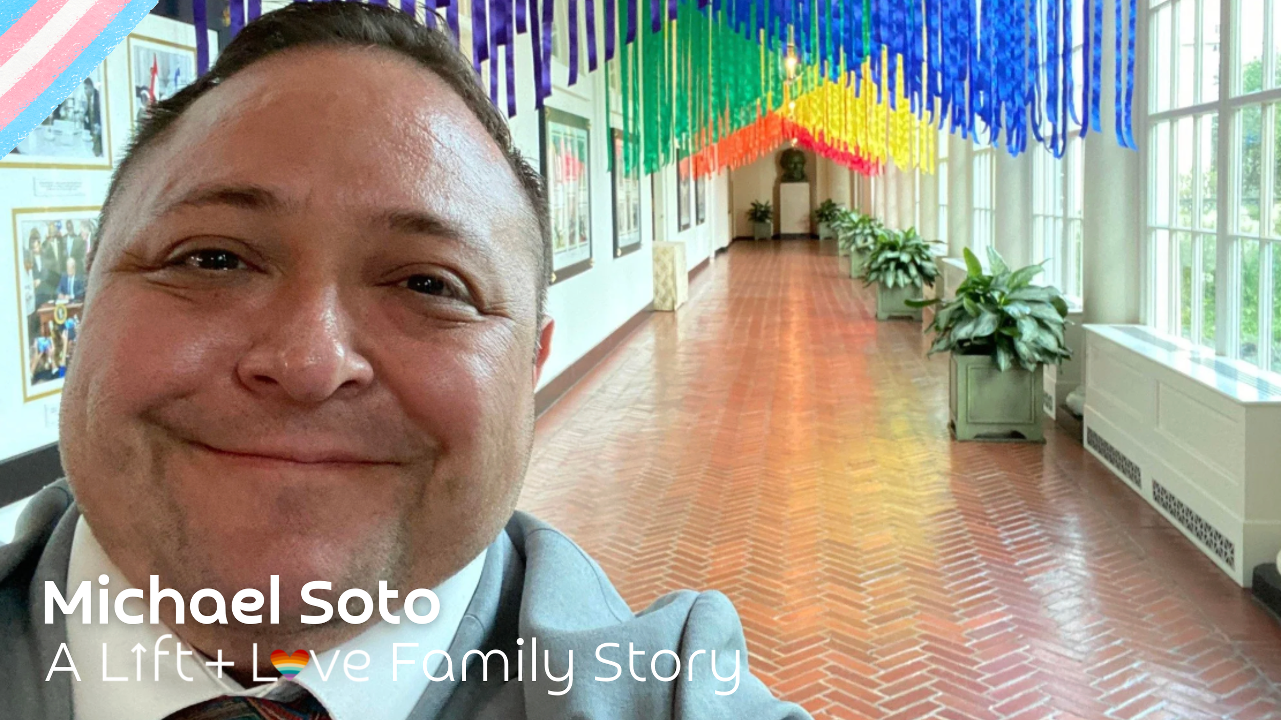 A man smiling in the foreground with colorful hanging decorations in a hallway with large windows and potted plants.