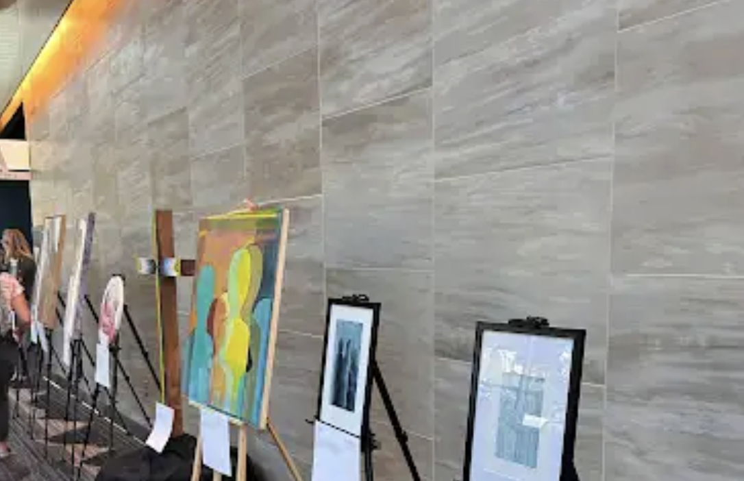 Indoor art exhibit with paintings on easels and framed artworks against a gray tiled wall.