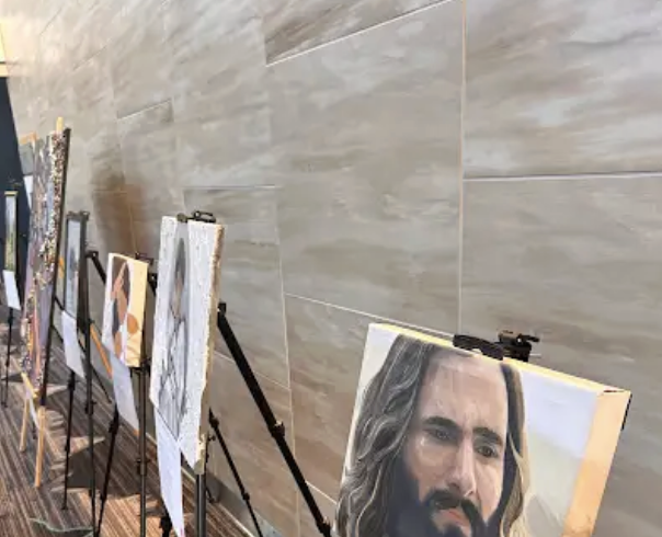 Multiple paintings displayed on easels in an art gallery, with a focus on a portrait of a man with long hair and a beard.