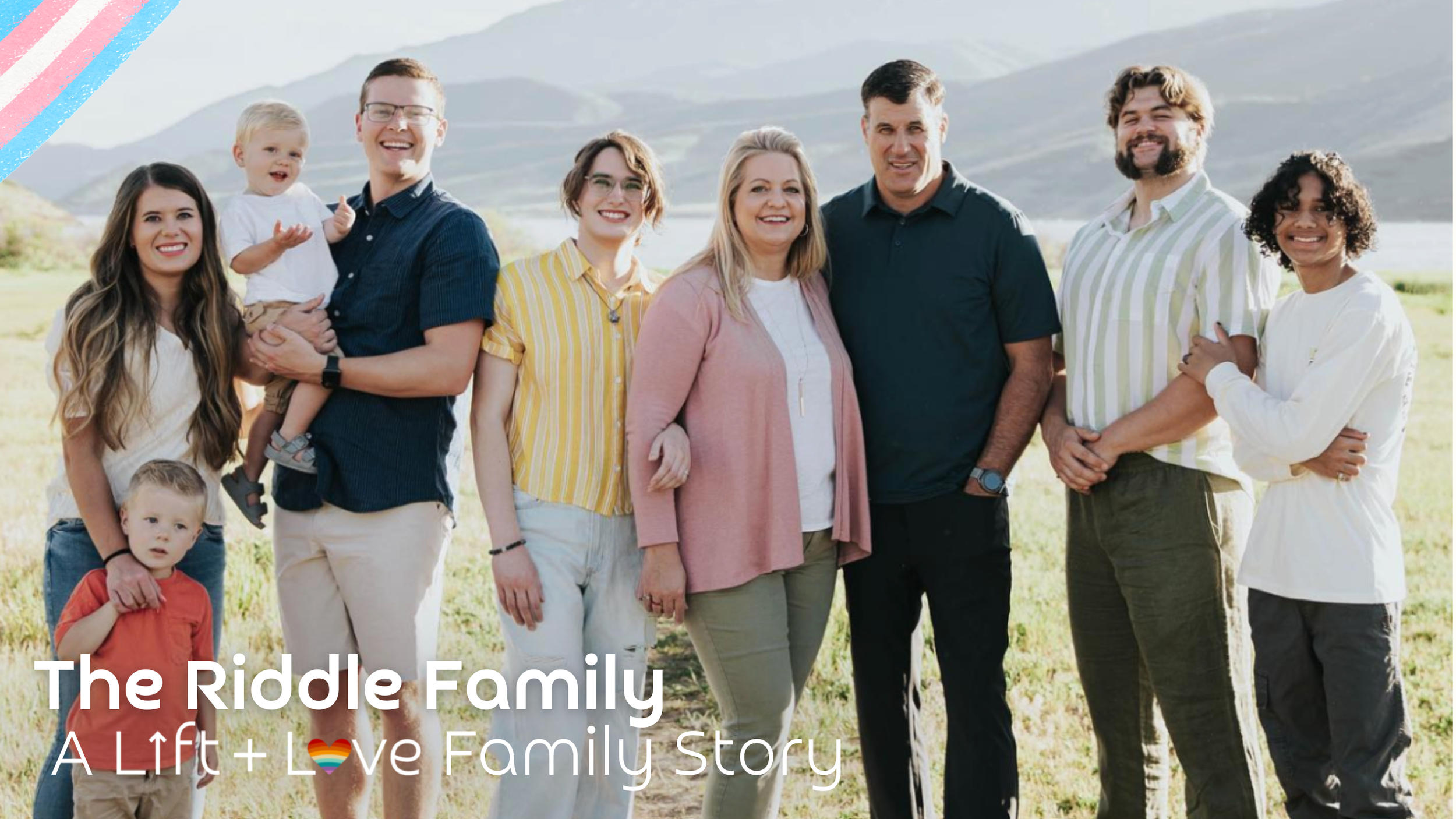 A multi-generational family standing outdoors on a grassy field with mountains in the background. The family includes adults and children smiling at the camera, dressed casually for a sunny day.