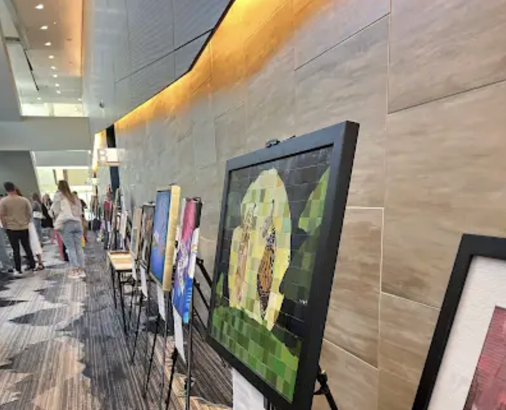 Art exhibition in a shopping mall with paintings on display on black easels, people walking and viewing art, interior with tile walls and ceiling lighting.