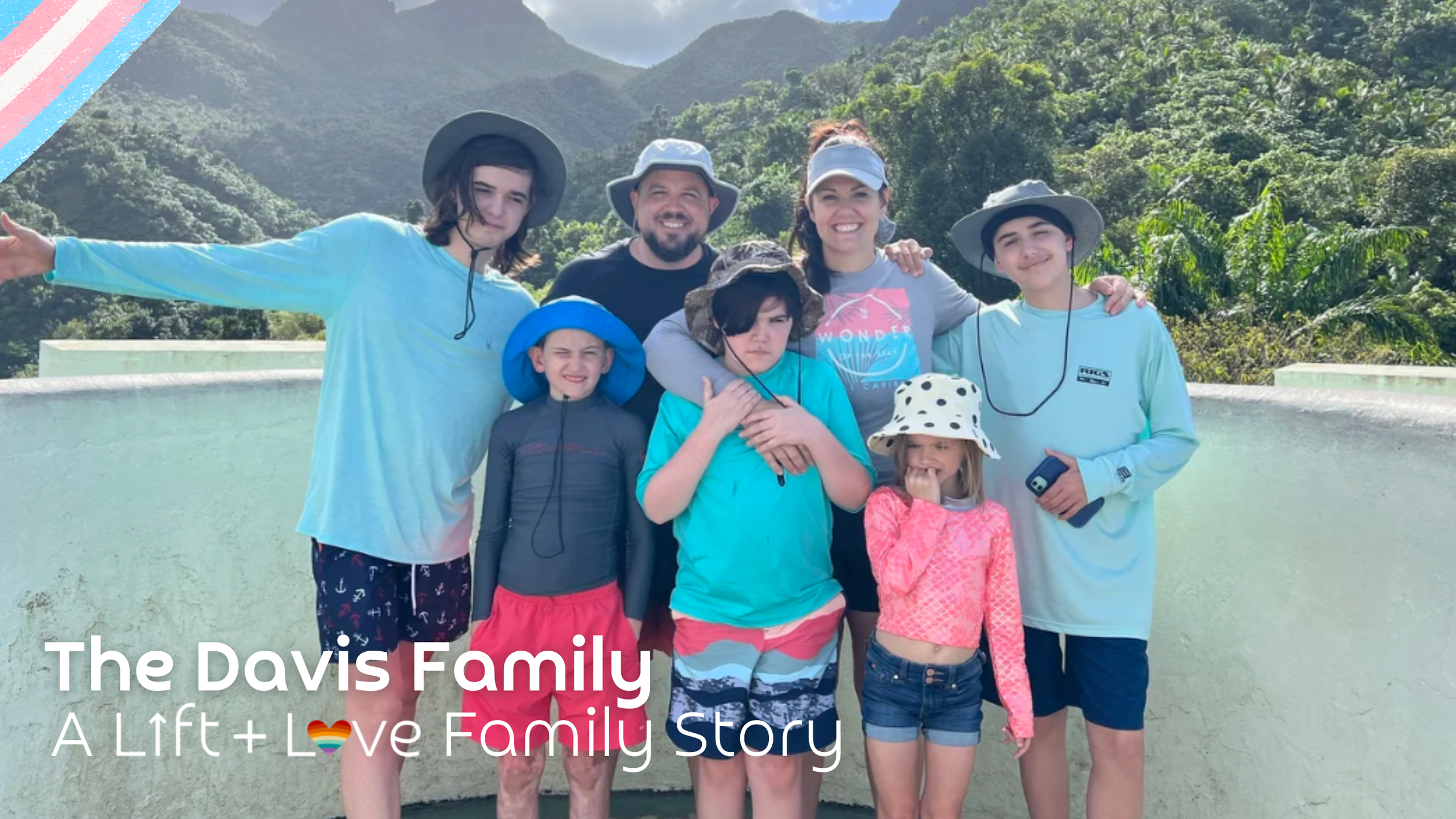 A smiling family of eight children and two adults standing outdoors with mountains and greenery in the background, wearing casual summer clothing and hats.
