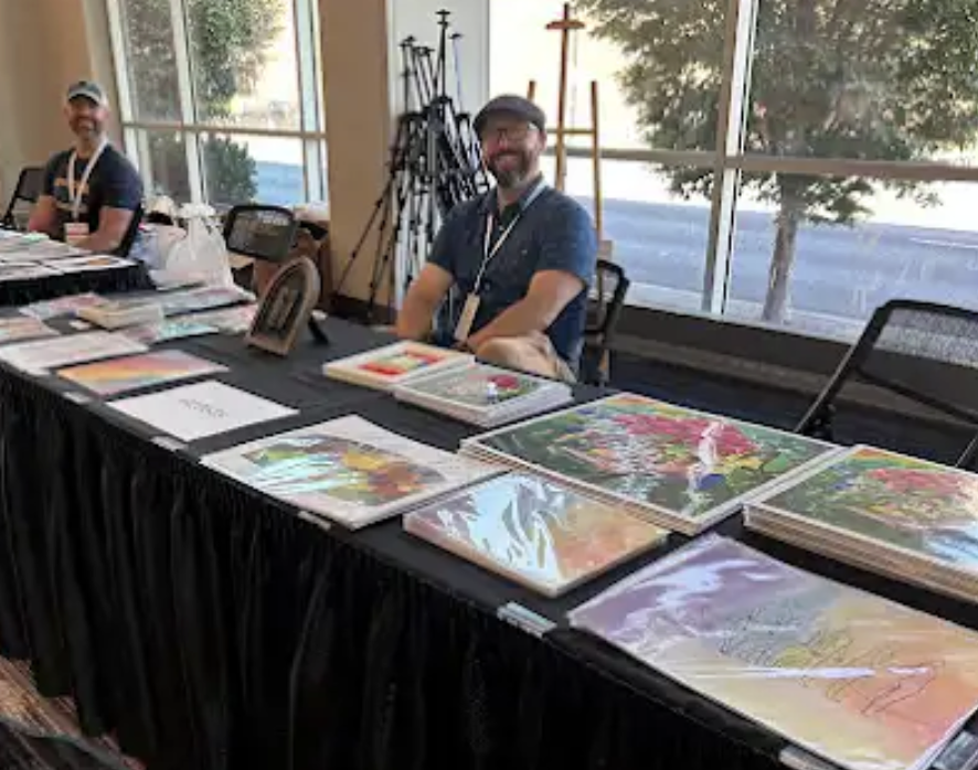 Two men seated at a table displaying colorful artwork, with art supplies and tripods behind them, in a room with large windows showing trees outside.