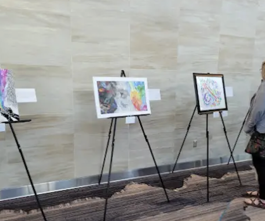 Three framed abstract artwork pieces displayed on black easels at an art exhibit.