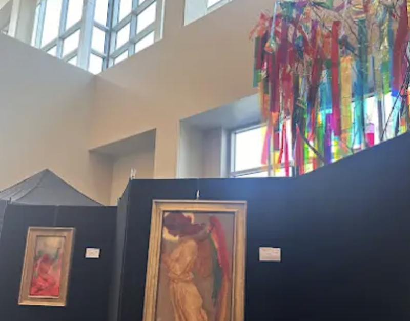 Art gallery display with framed paintings on black panels and colorful hanging decorations in a bright room with large windows.