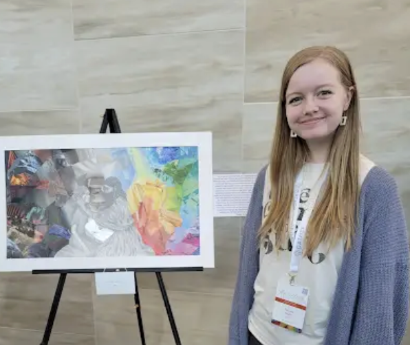 Young woman with long red hair standing next to colorful artwork on display at an art exhibit.