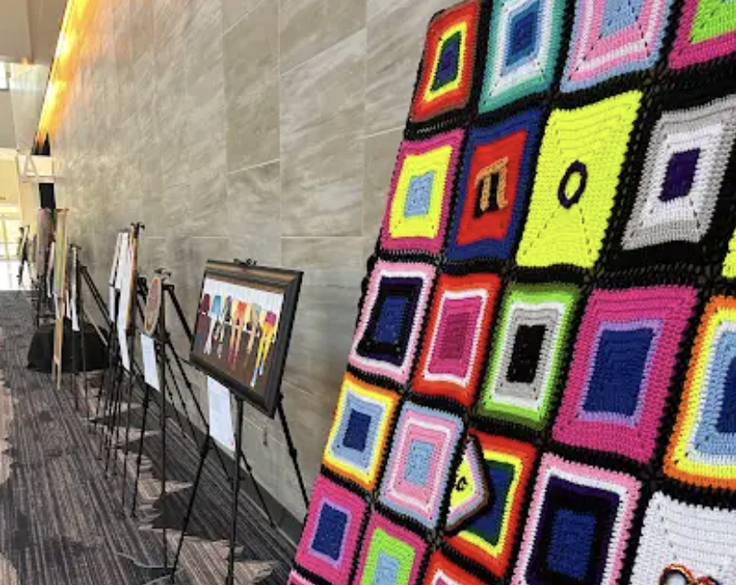 Colorful artwork on display, including framed paintings and a large crocheted piece with multicolored square patterns, exhibited on easels and a wall in what appears to be an art gallery or exhibition space.
