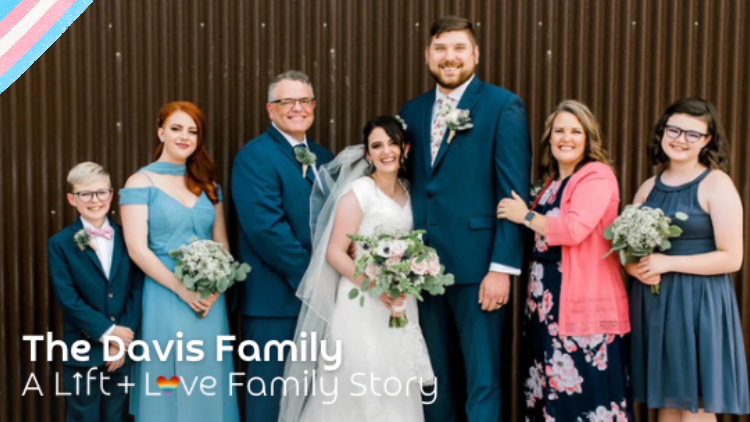 Group of eight people, including bride and groom, smiling and posing for a photo at a wedding reception, with a wooden wall background.