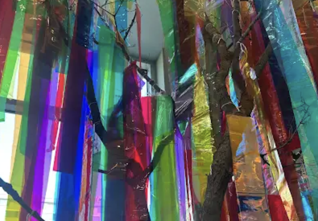 Colorful hanging fabric strips in various bright colors like green, pink, yellow, blue, and orange, with a central tree branch and some smaller branches inside a room.