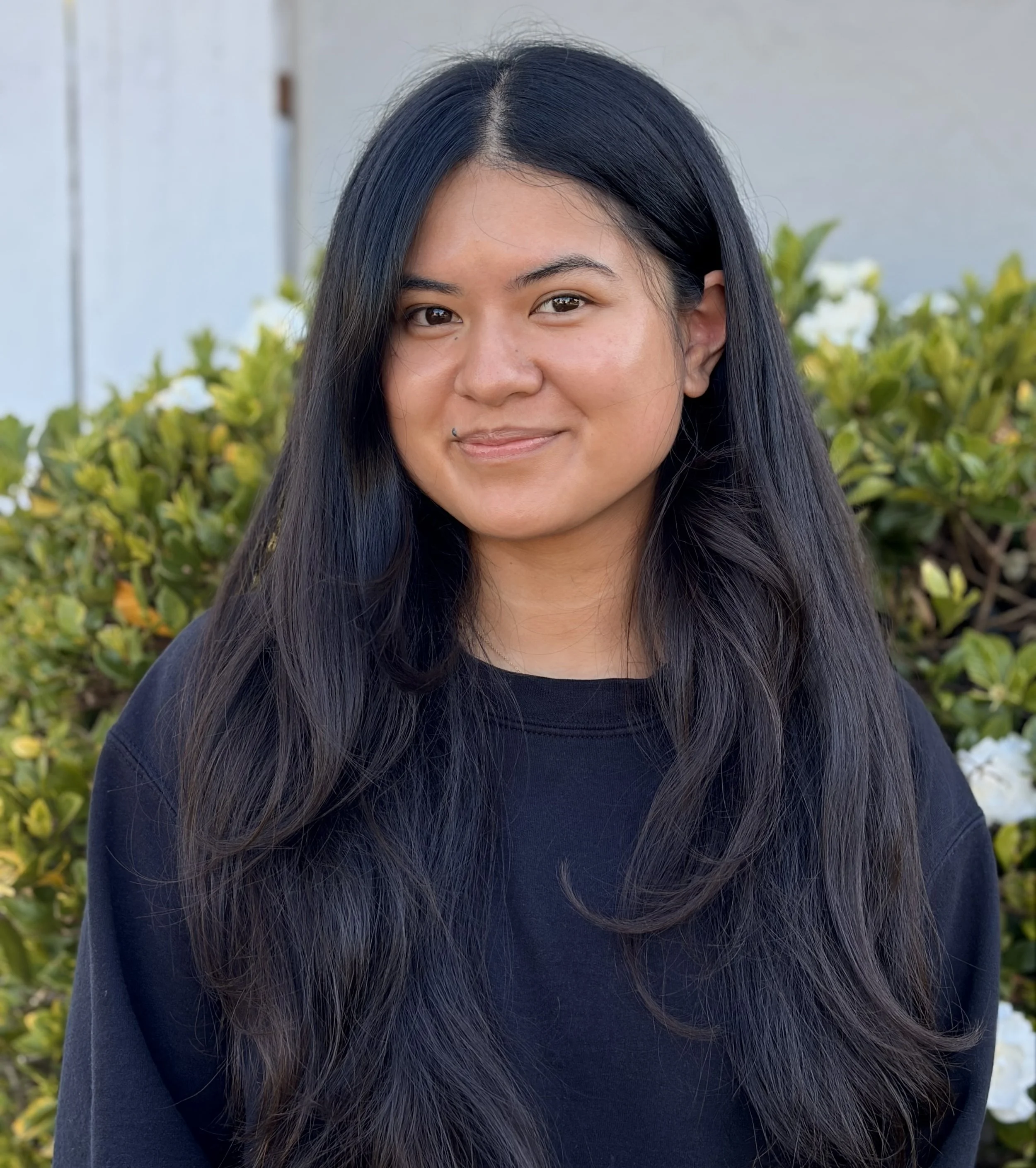 Photo of Caroline outdoors with a flower bush behind her. Caroline is a young Asian woman with long black hair cascading down her shoulders with a closed mouth smile