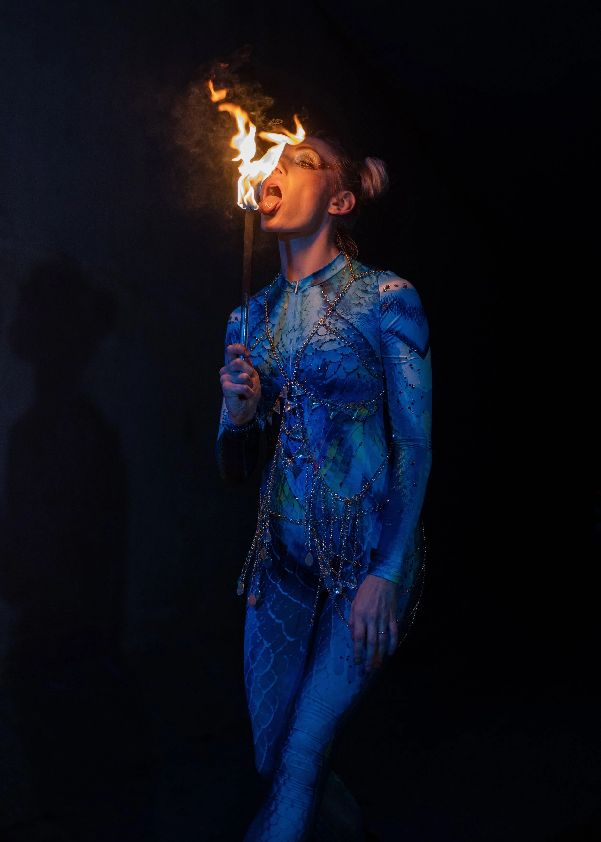 A person dressed in a shiny, blue, patterned costume holding a lit torch with flames, standing against a dark background, appearing to perform in a fire act.