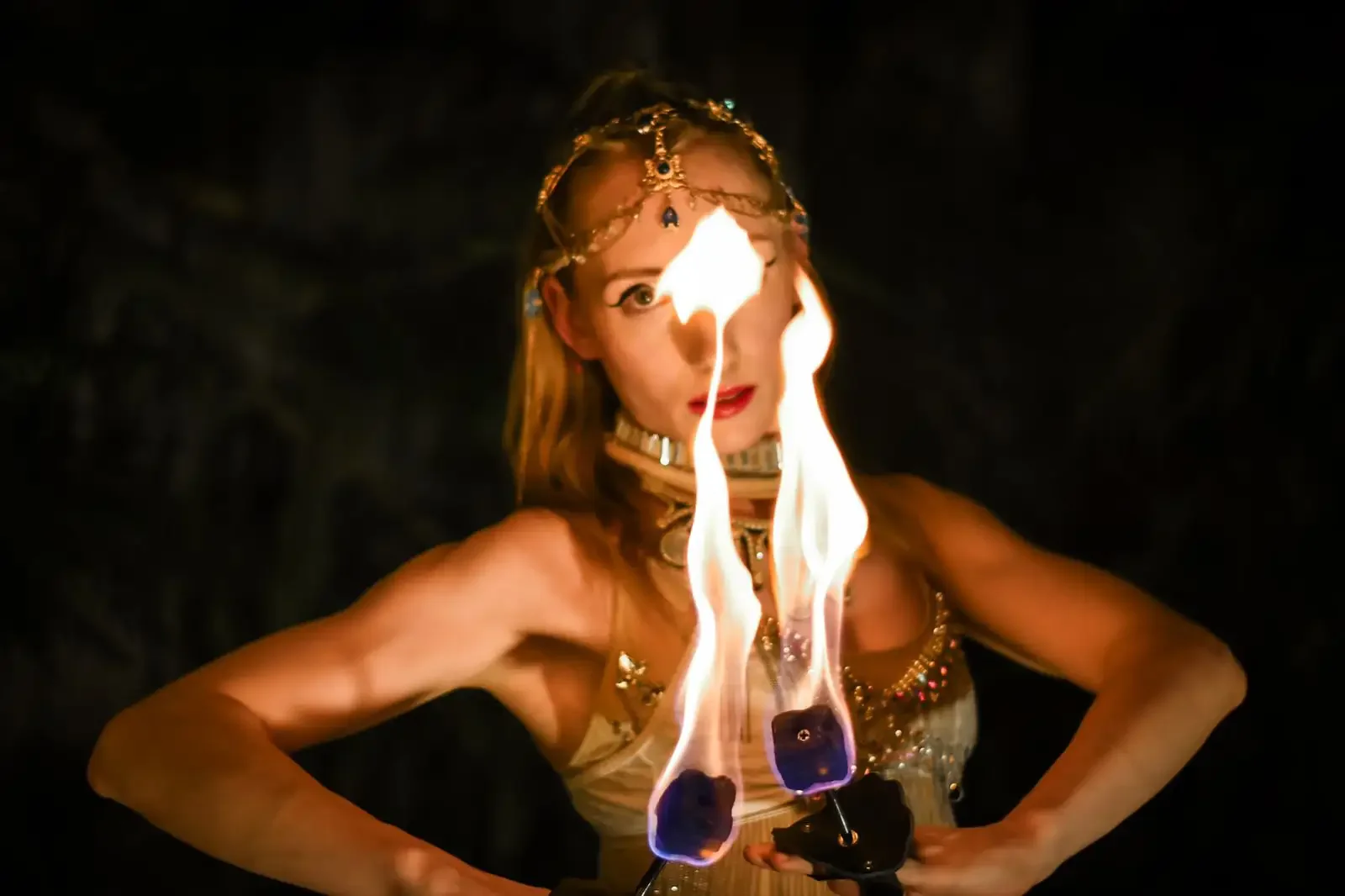 A woman with long hair, wearing jewelry and makeup, holding a flaming torch with fire in front of her face against a dark background.