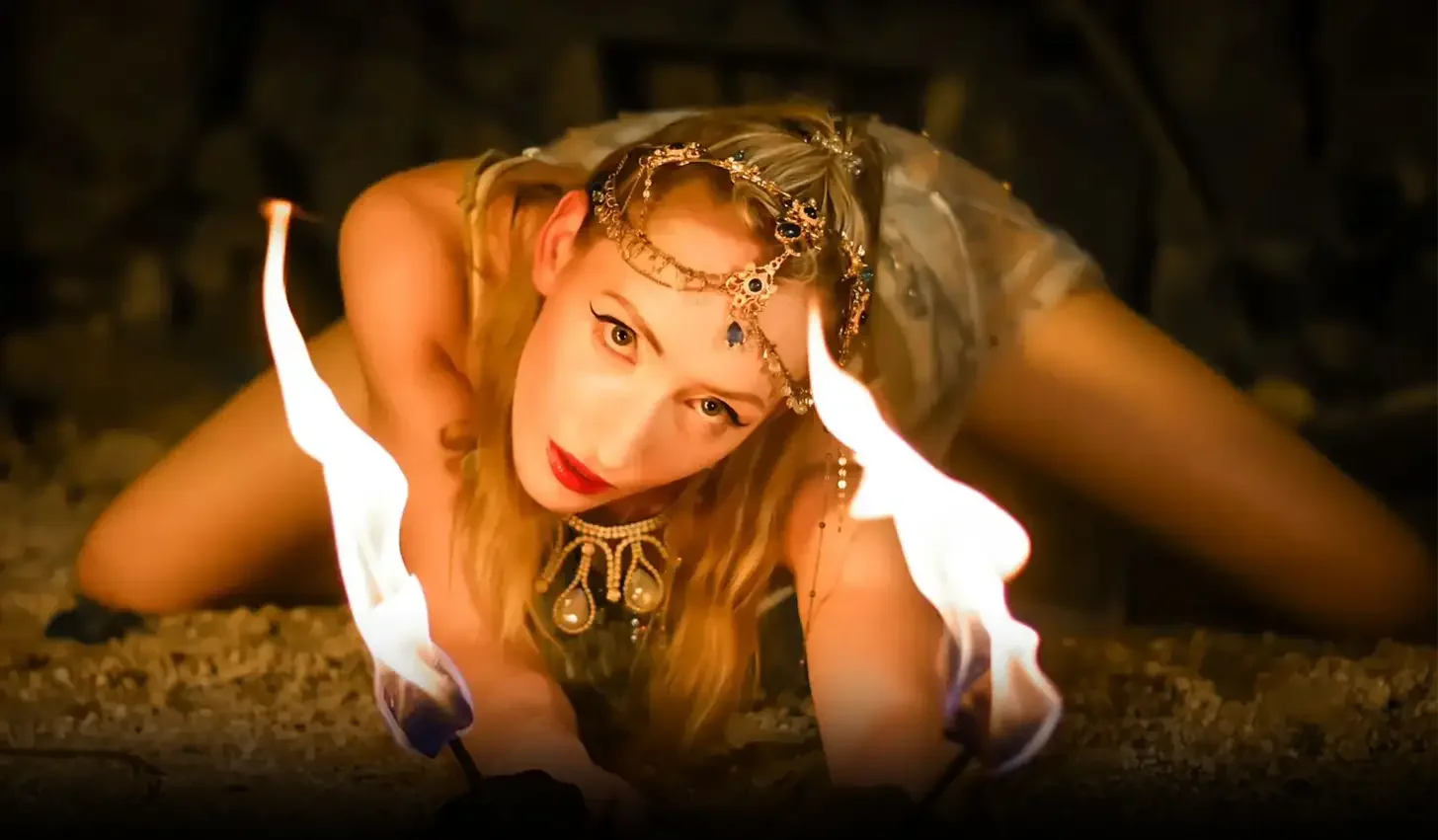 A woman dressed as a demon or mythical creature crawling on the ground with flames on her hands, wearing jewelry and makeup, with a serious expression.