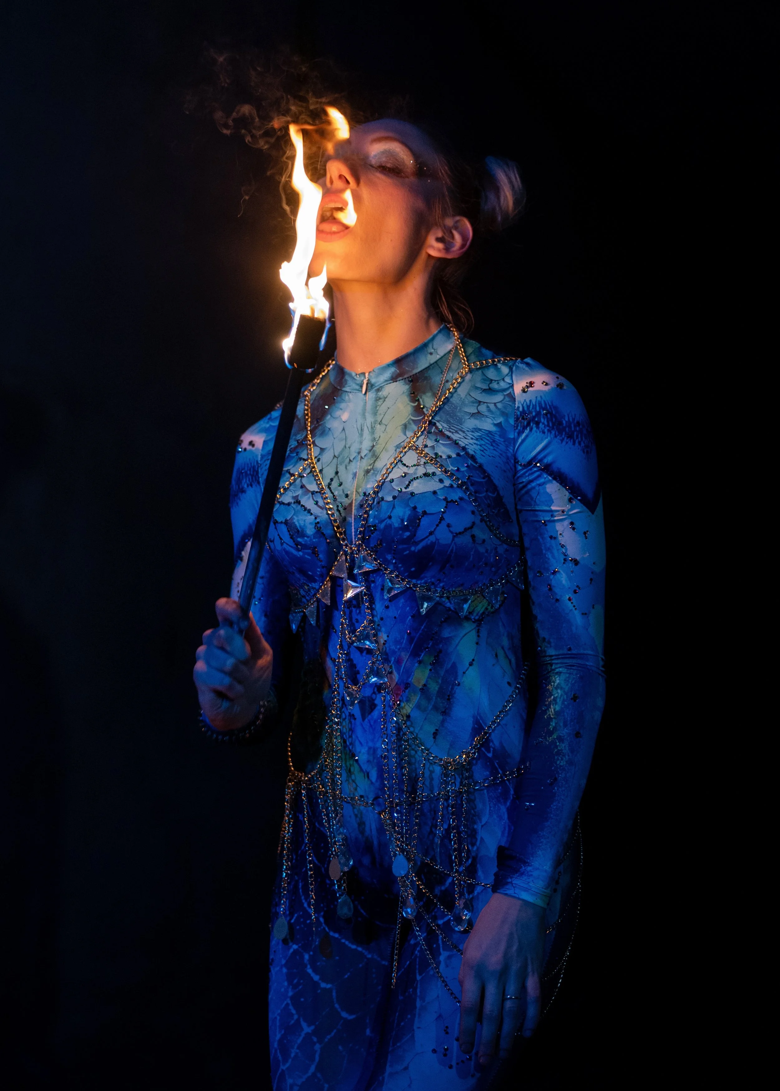 A person in a blue patterned outfit is performing a fire-eating act, holding a flaming torch close to their face in a dark setting.