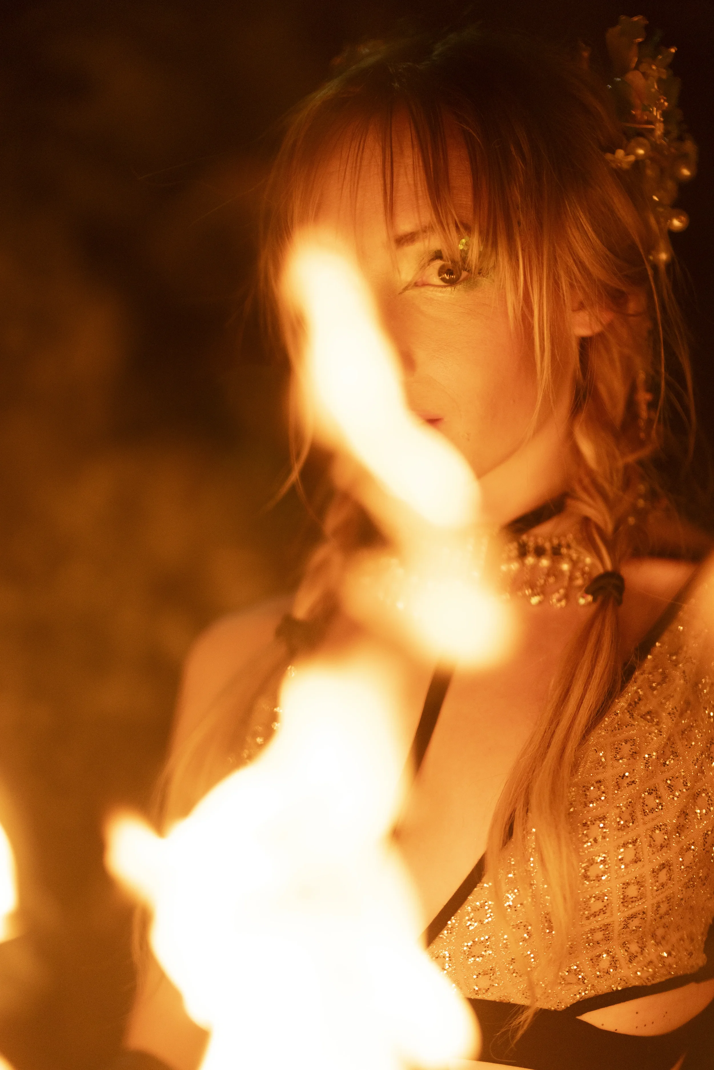 A woman with blonde hair styled in braids and flowers, wearing a sparkly dress and jewelry, is partially obscured by flames in a dimly lit setting.