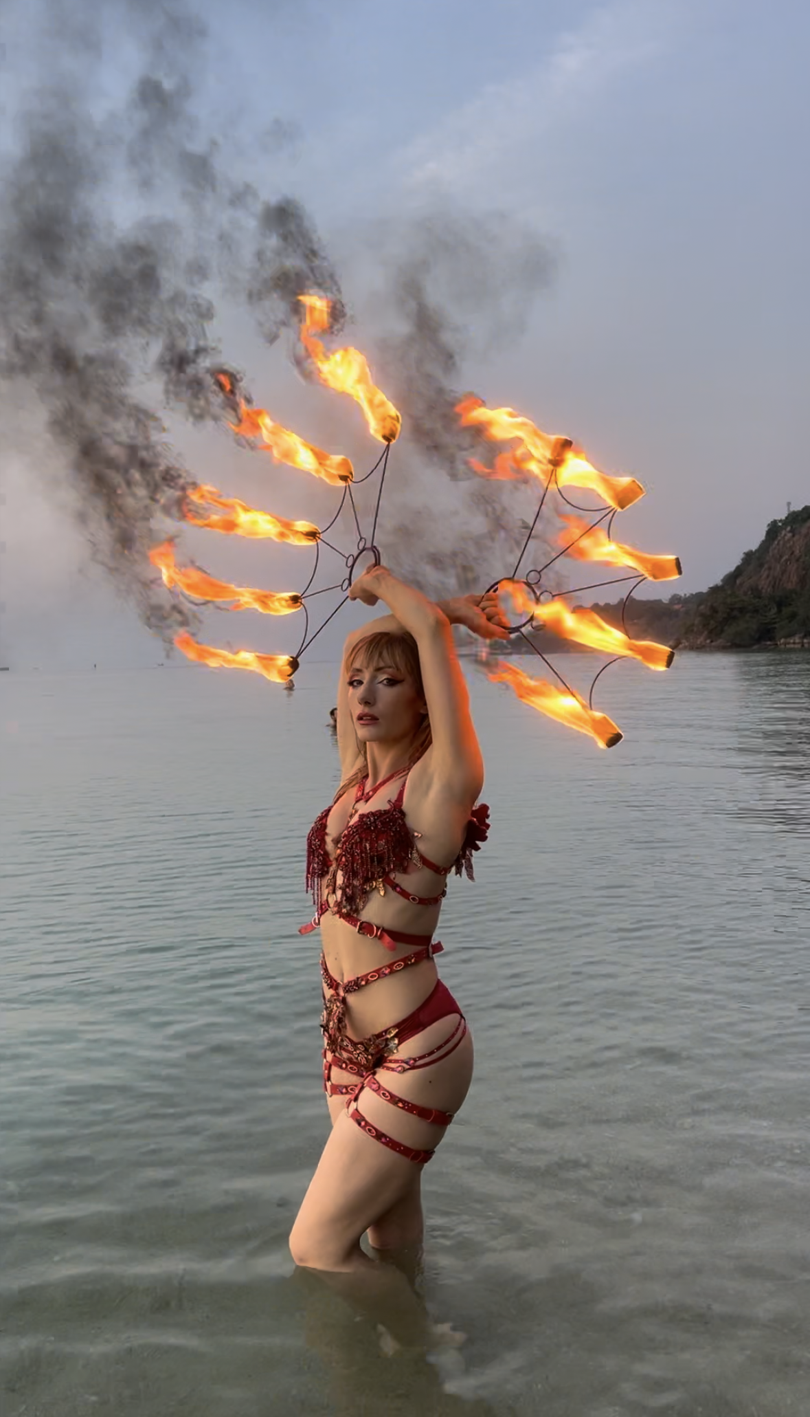 A woman holding fire torches in both hands above her head, standing in water with a scenic landscape in the background.