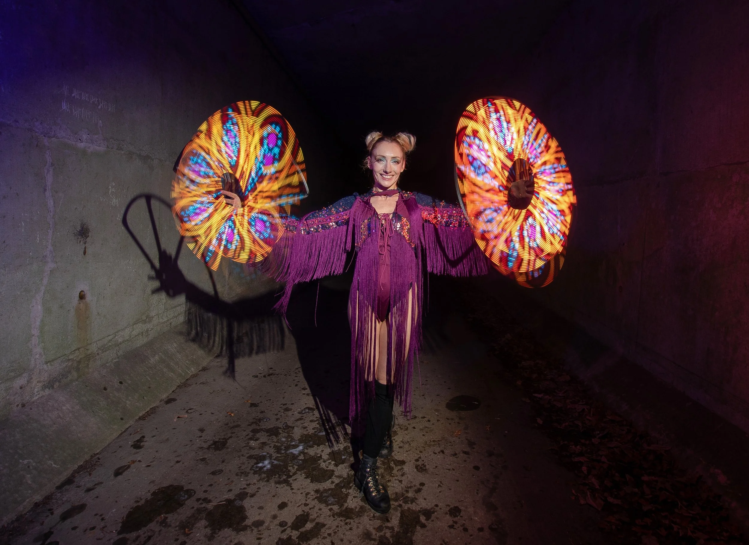 A woman in a purple sequined fringed outfit with two large LED fans, posing in a dark tunnel.