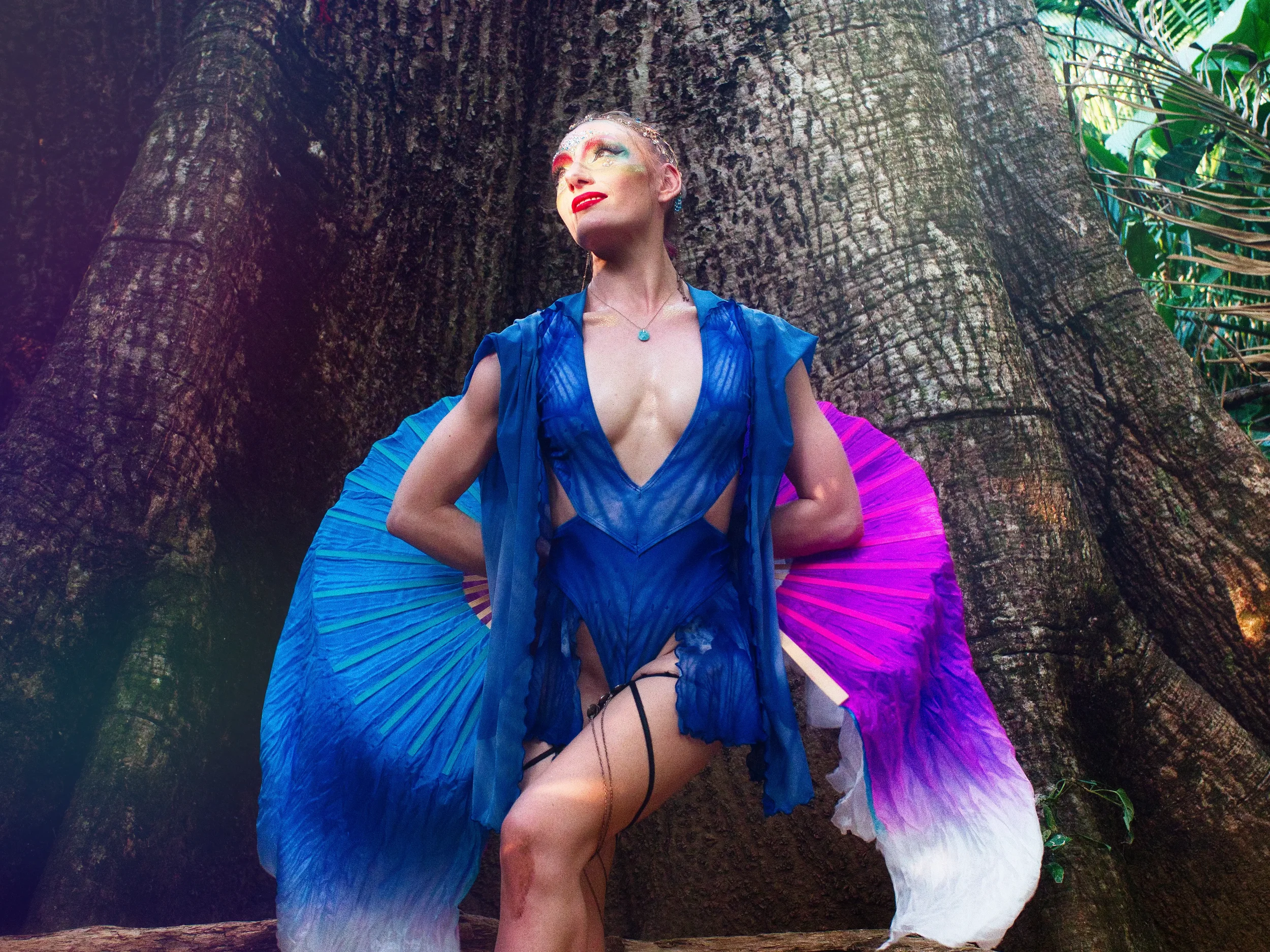 a woman in a blue outfit poses against a large tree with green foliage in the background.