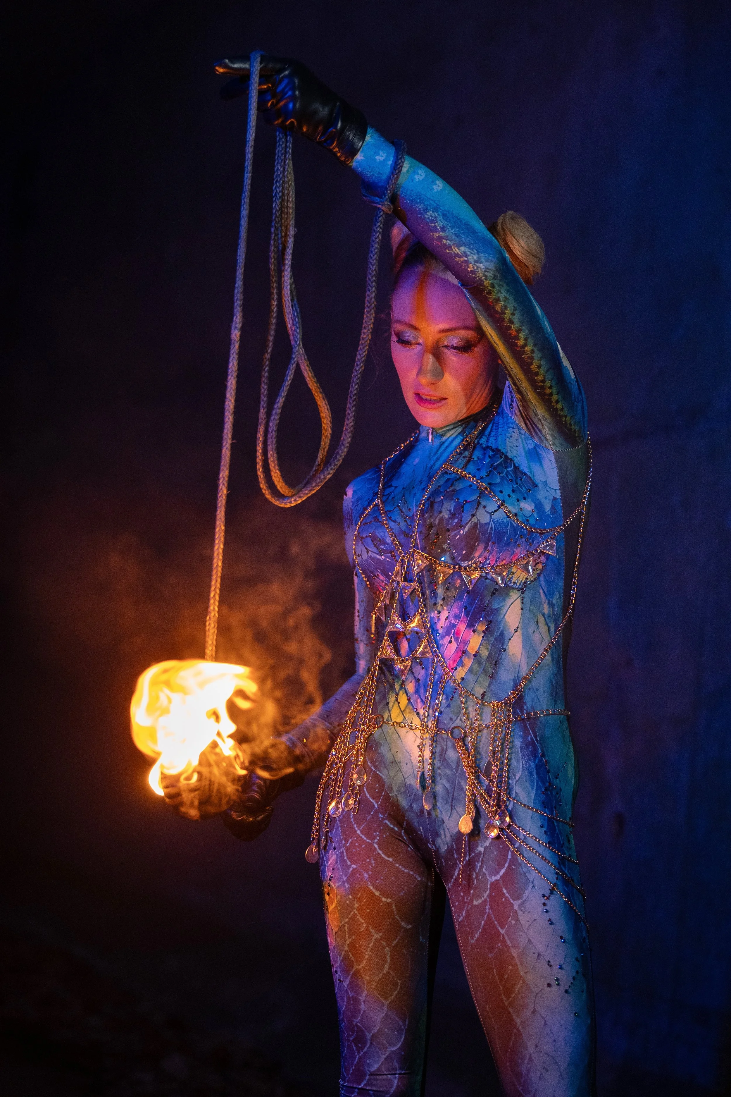 A woman dressed in an elaborate, colorful costume with gold chains, holding a flaming torch in her gloved hand. The background is dark, highlighting the flame and her costume.